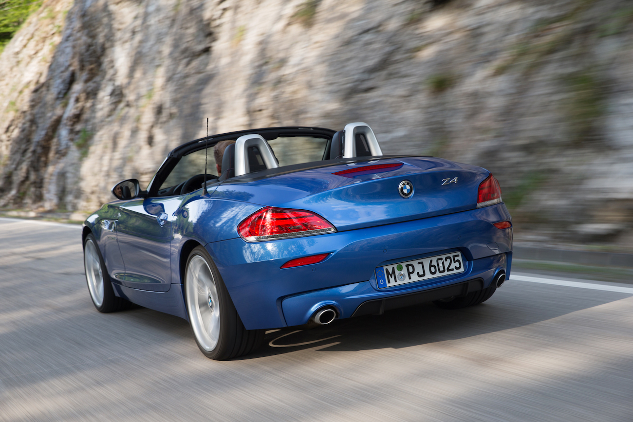 Blauer BMW Z4 Roadster fährt auf einer kurvigen Straße entlang einer Felswand, Verdeck geöffnet. Dynamische Fahrt bei Tageslicht.