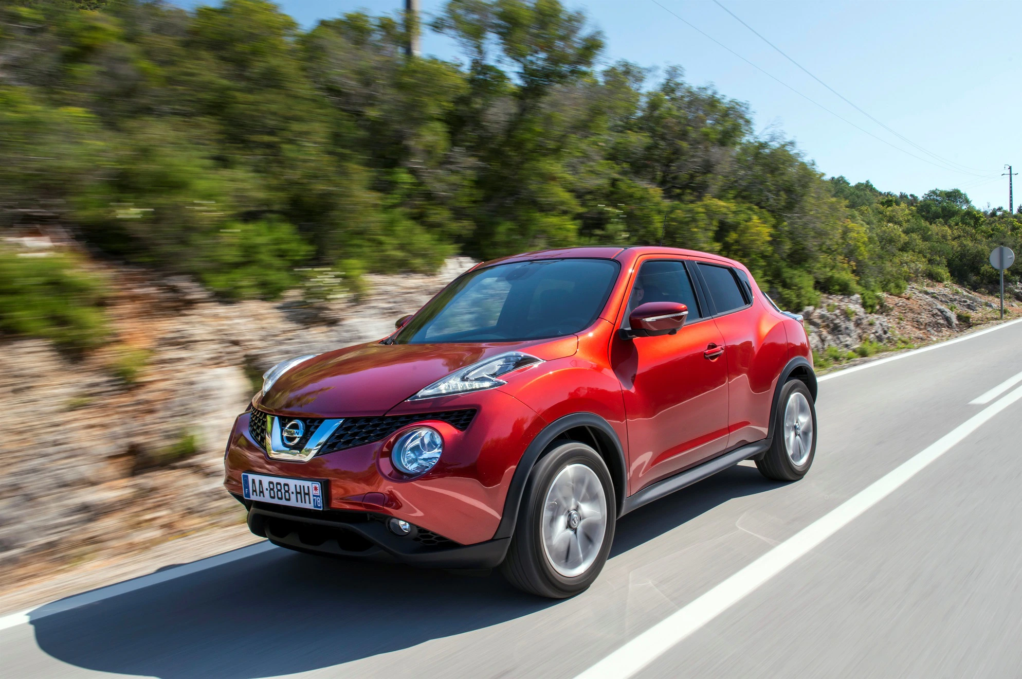 Roter Nissan Juke fährt auf einer Landstraße, umgeben von Bäumen und Hügeln. Der Himmel ist klar und sonnig.