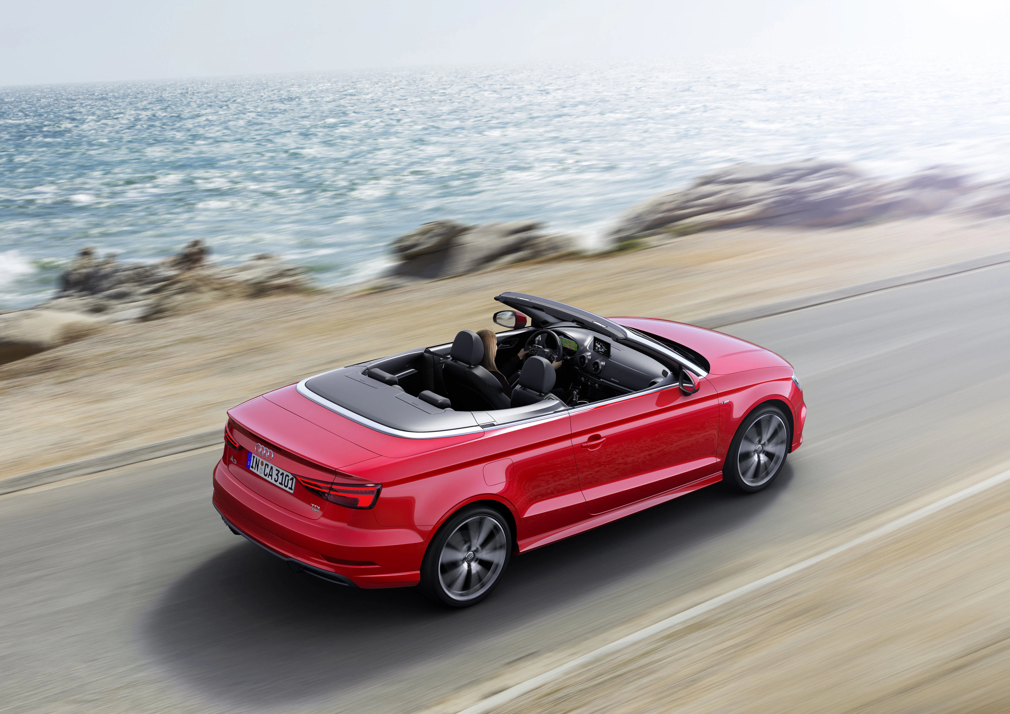 Rotes Cabrio fährt auf einer Küstenstraße entlang des Meeres bei sonnigem Wetter. Das Verdeck ist geöffnet.