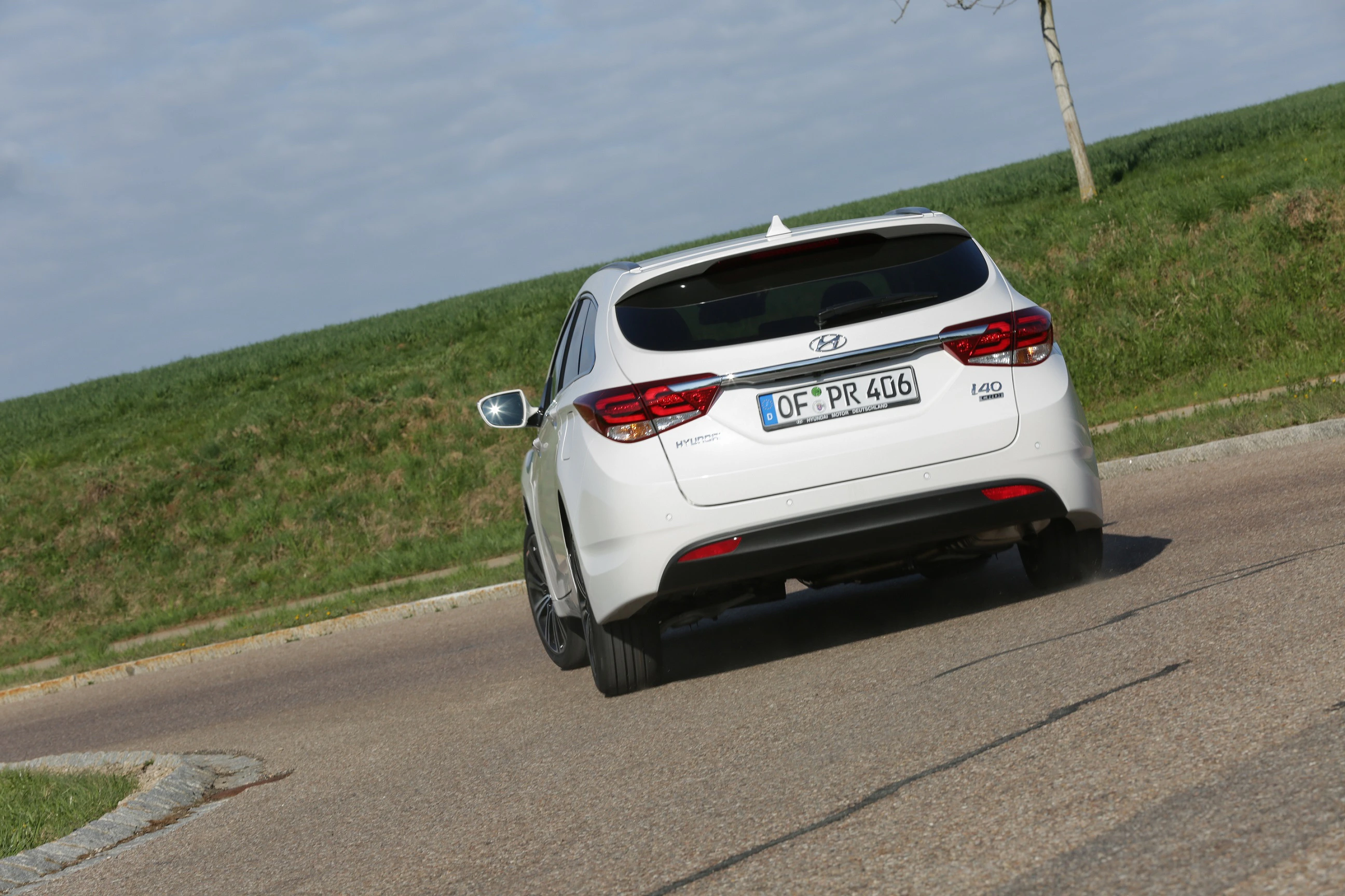 Weißer Hyundai i40 Kombi fährt auf einer ländlichen Straße bei bewölktem Himmel. Im Hintergrund grünes Gras und blauer Himmel.