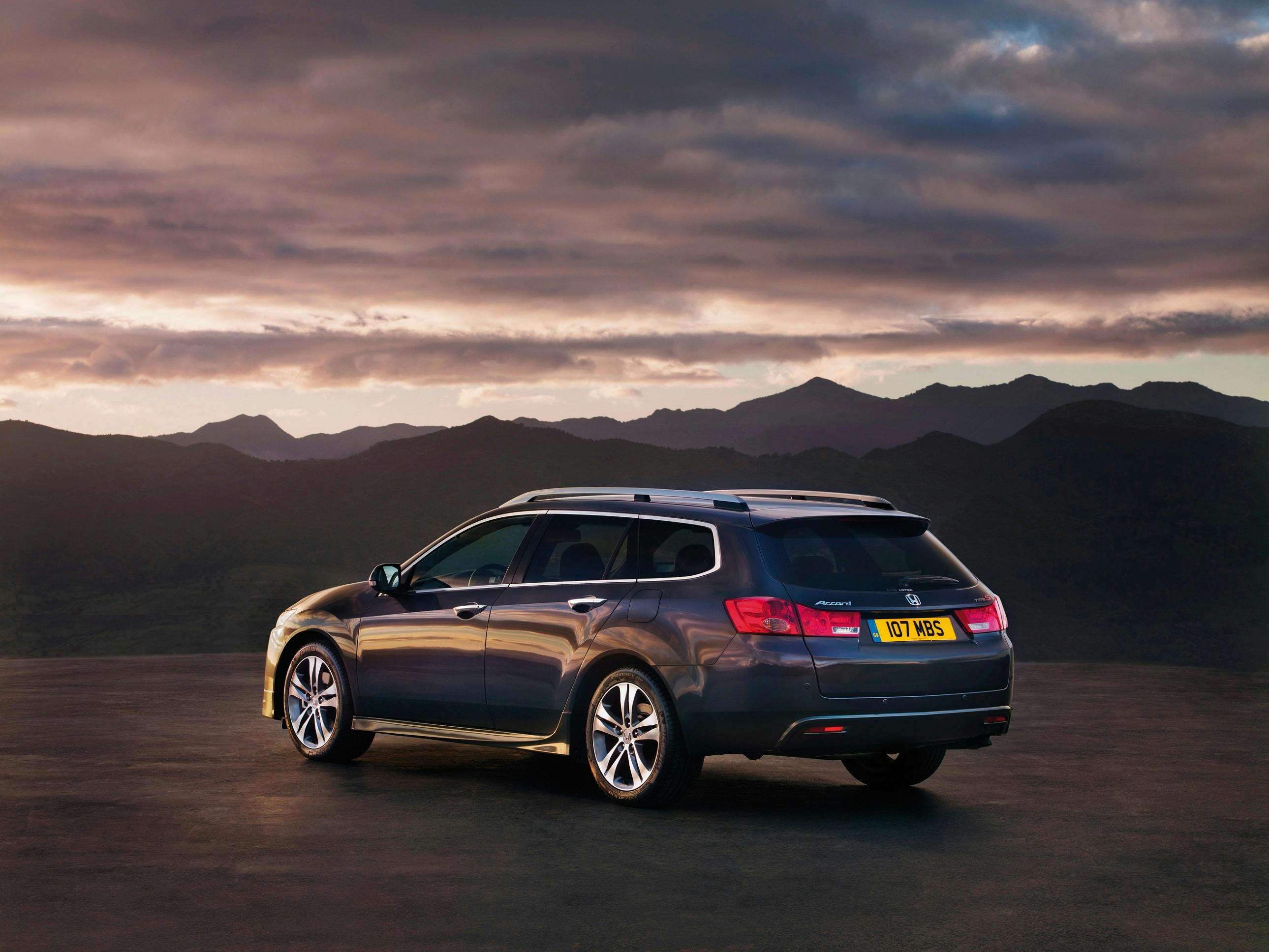 Dunkelgrauer Kombi auf einer Straße vor einer Bergkulisse bei Sonnenuntergang. Der Himmel ist dramatisch bewölkt.