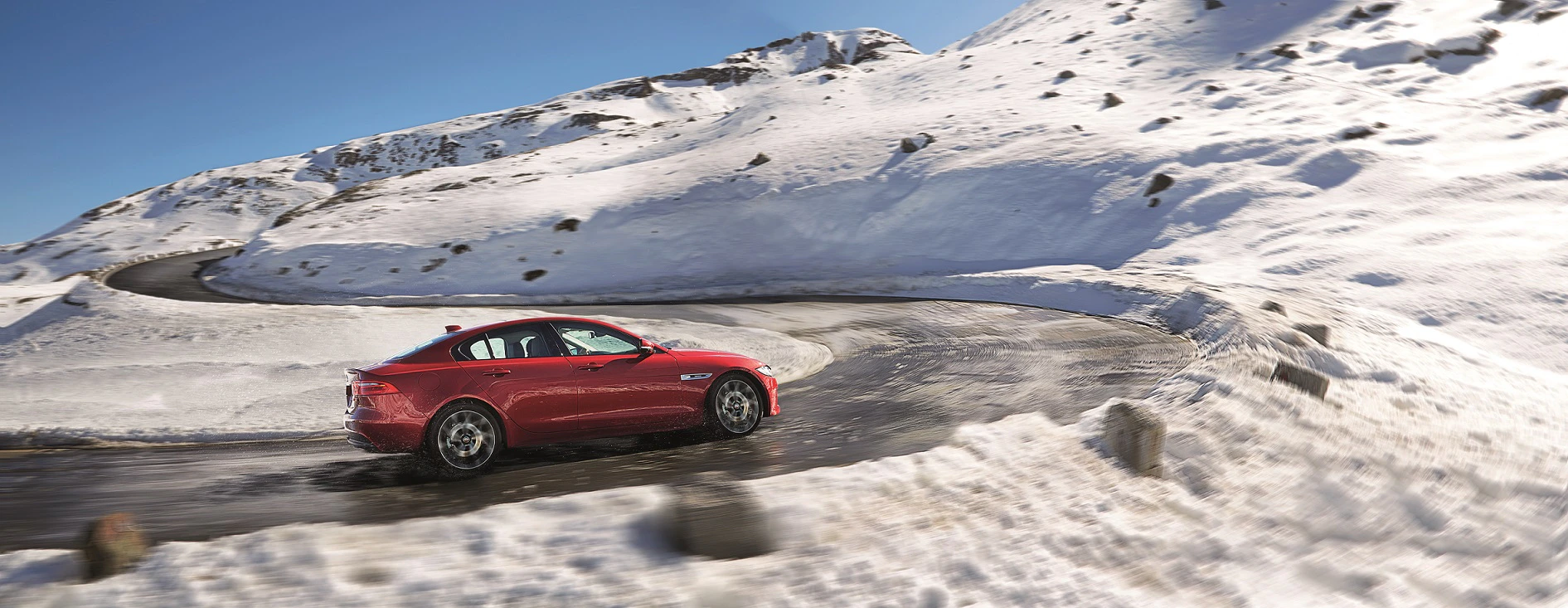 Rotes Auto fährt auf kurviger, schneebedeckter Bergstraße unter klarem, blauem Himmel. Schnee bedeckt die umliegende Landschaft.