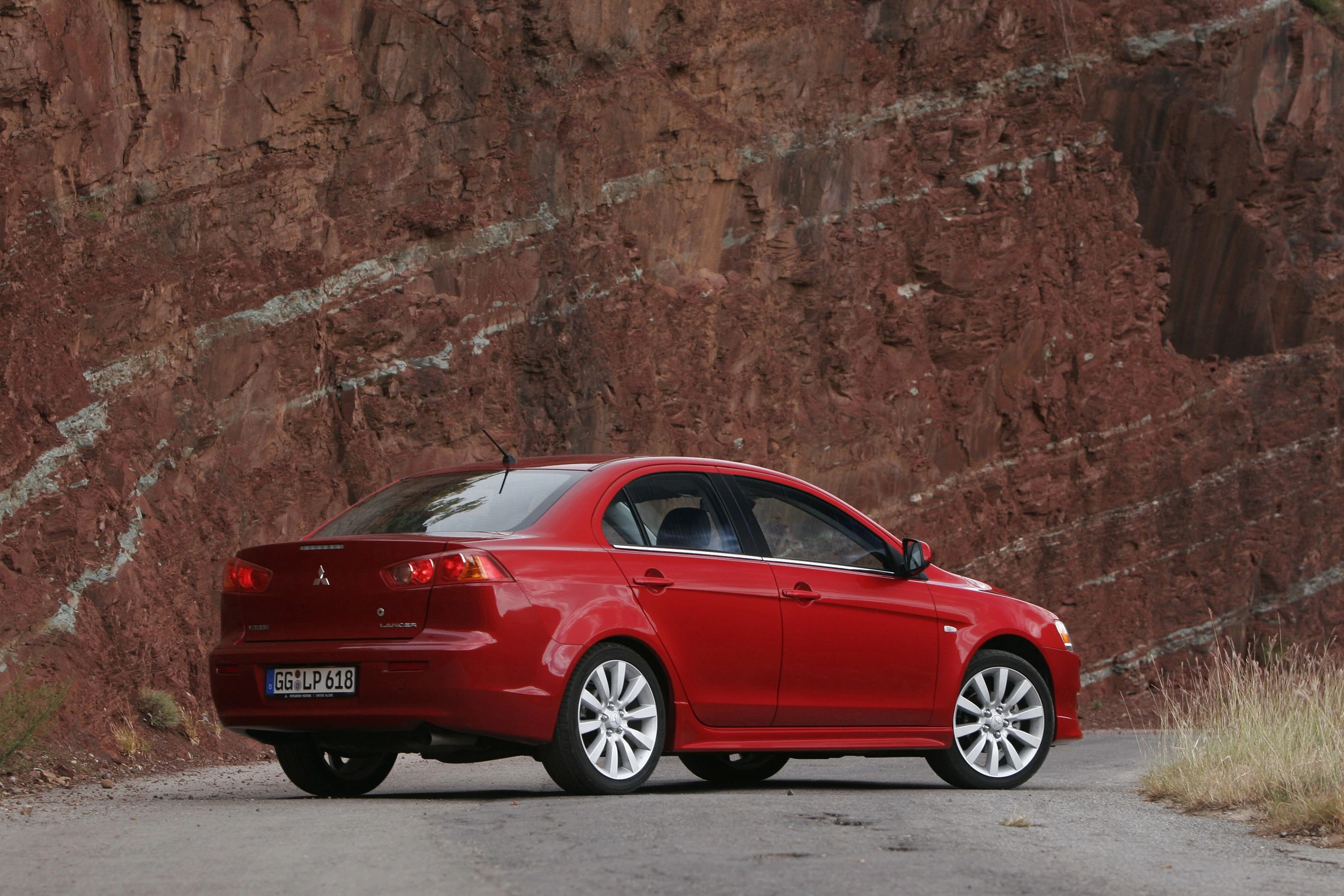 Roter Mitsubishi Lancer auf einer Straße in einer felsigen Landschaft, Heckansicht, mit markanten Felsformationen im Hintergrund.