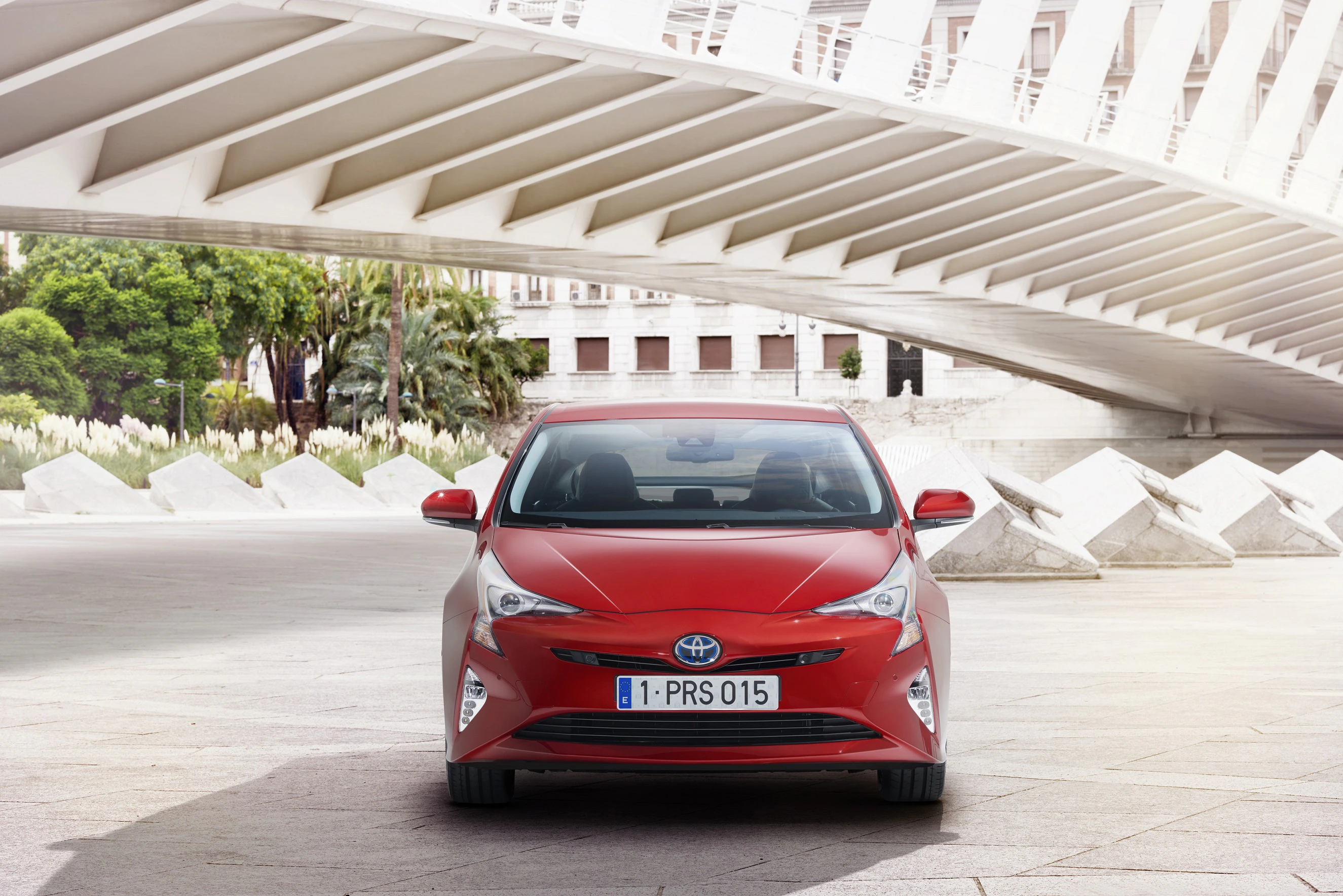 Roter Toyota Prius steht unter modernem, geschwungenem Brückengewölbe, umgeben von urbaner Architektur und grünen Pflanzen.
