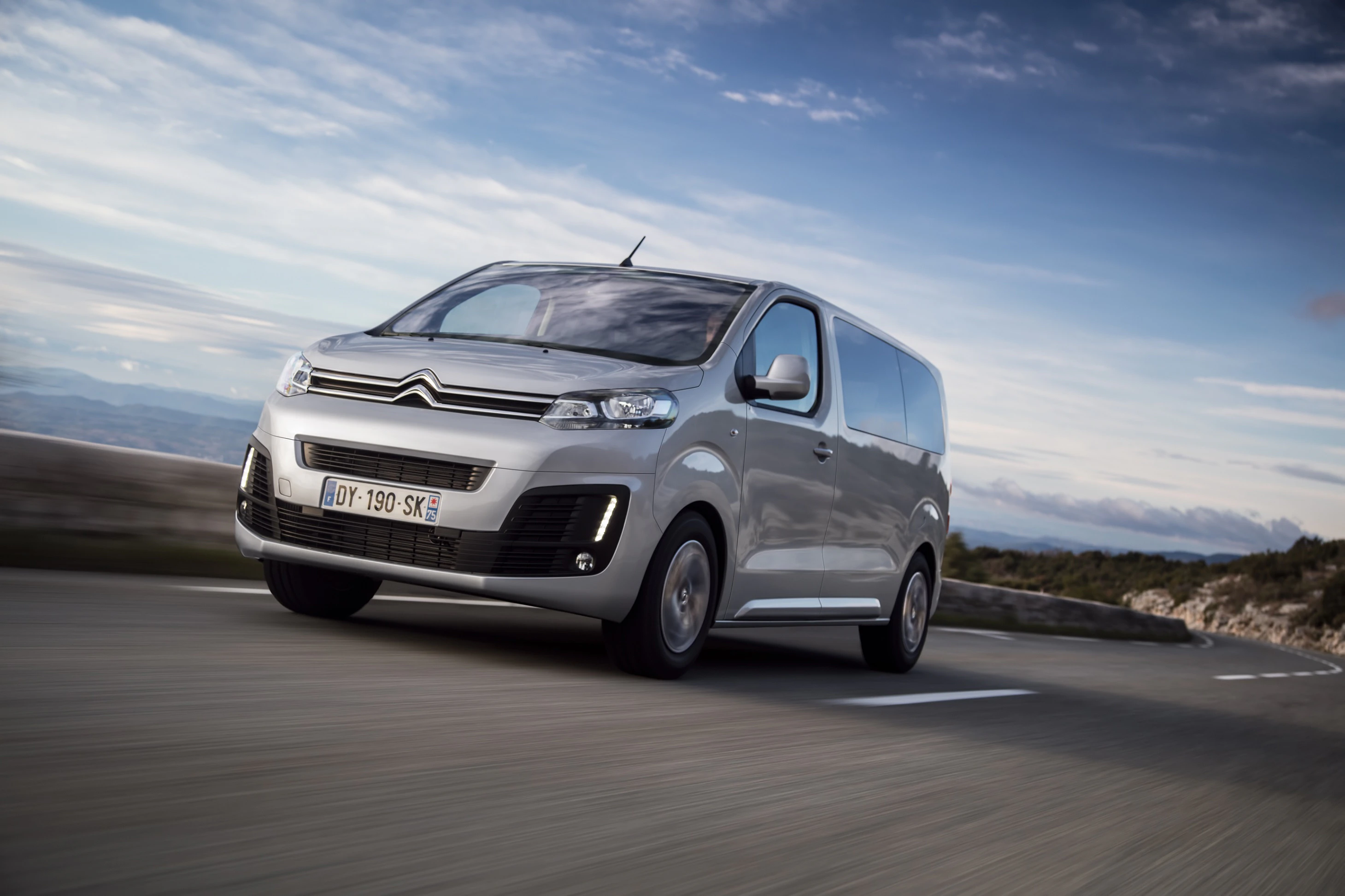 Grauer Citroën Van fährt auf einer Landstraße bei bewölktem Himmel. Die Straße verläuft durch eine hügelige Landschaft.