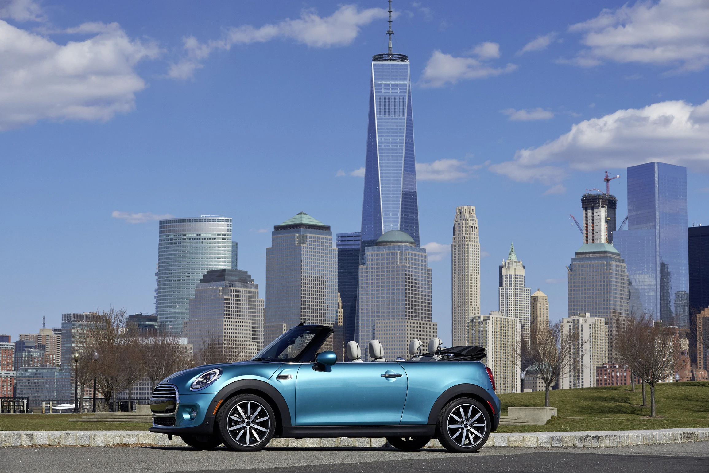 Blauer Mini Cooper Cabrio vor der Skyline von New York City mit dem One World Trade Center im Hintergrund an einem klaren Tag.