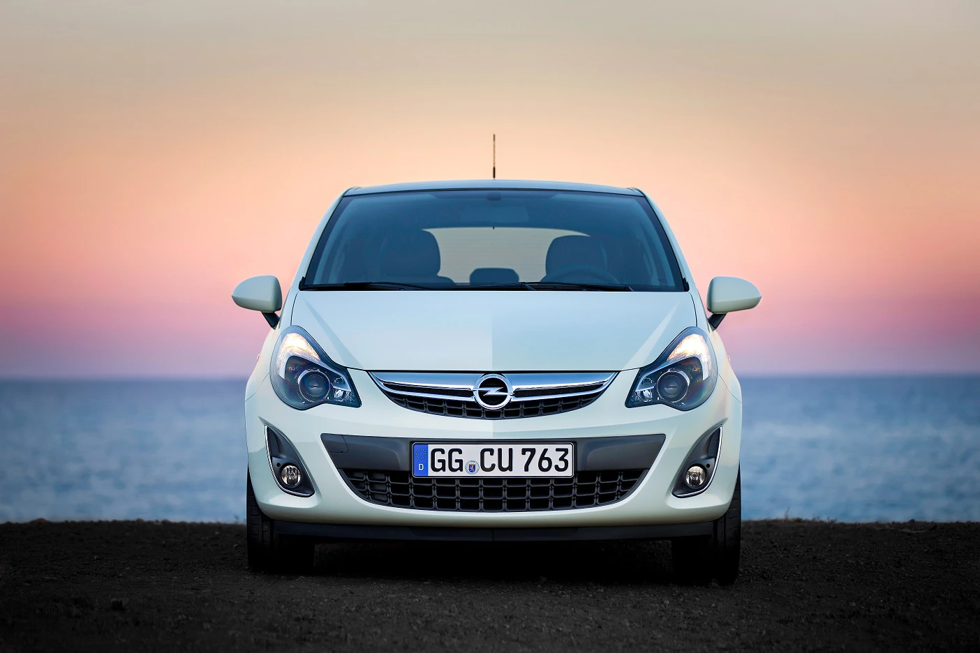 Weißer Opel Corsa steht vor einem farbenfrohen Sonnenuntergang am Meer.