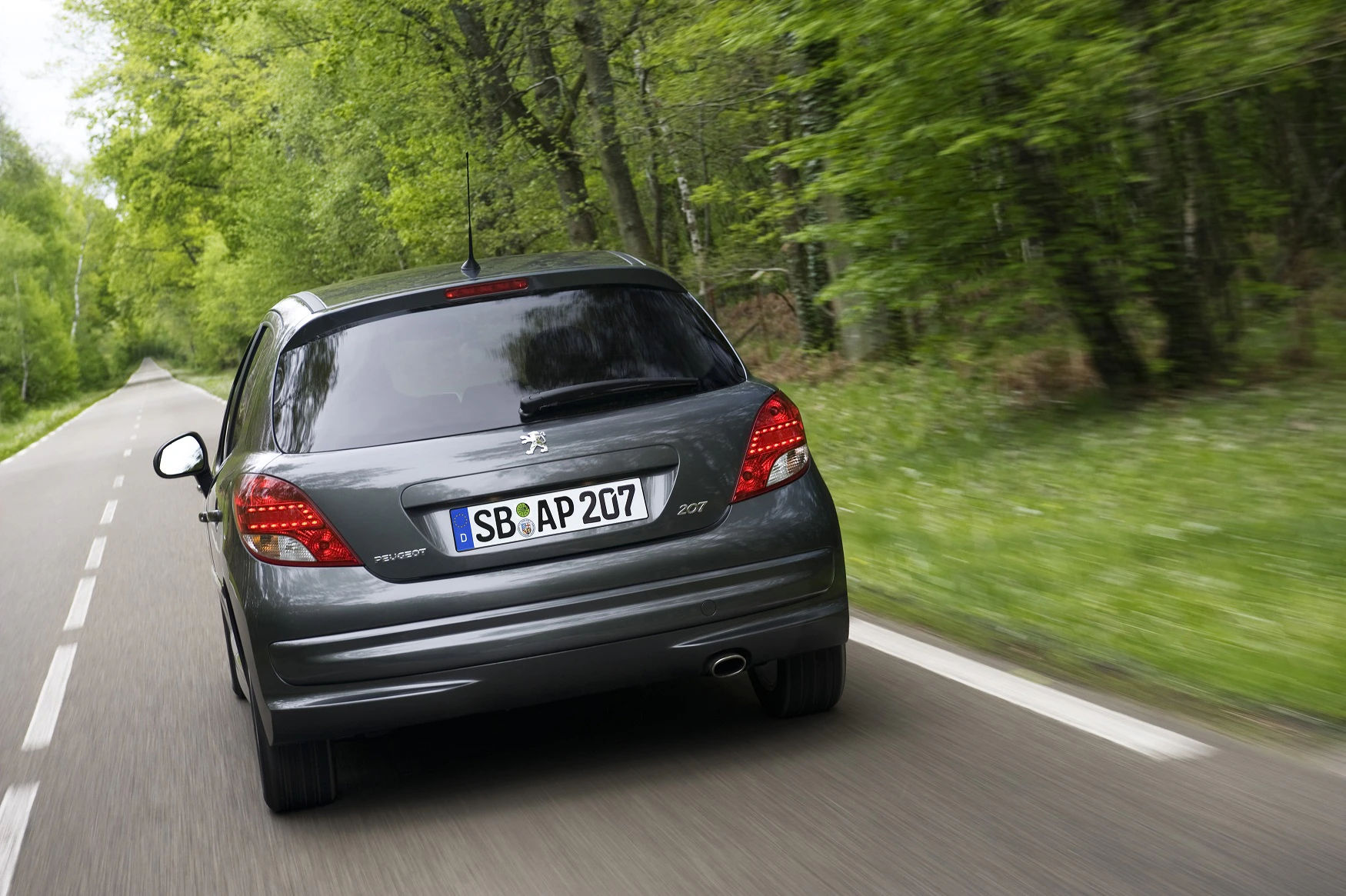 Silbergrauer Peugeot 207 fährt auf einer ländlichen Straße, umgeben von grünen Bäumen. Sicht von hinten.