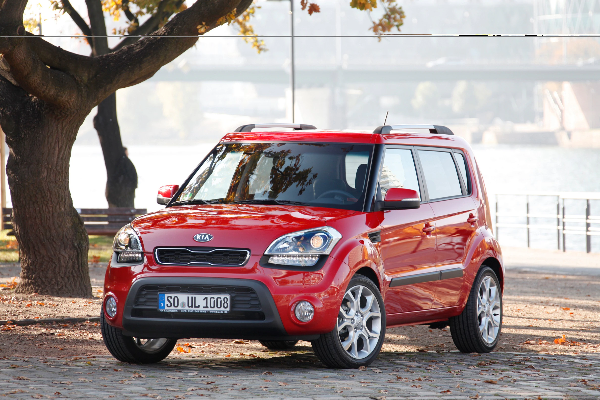 Roter Kia Soul parkt auf einem Kopfsteinpflaster vor herbstlichen Bäumen, im Hintergrund ist ein Fluss und eine Brücke zu sehen.