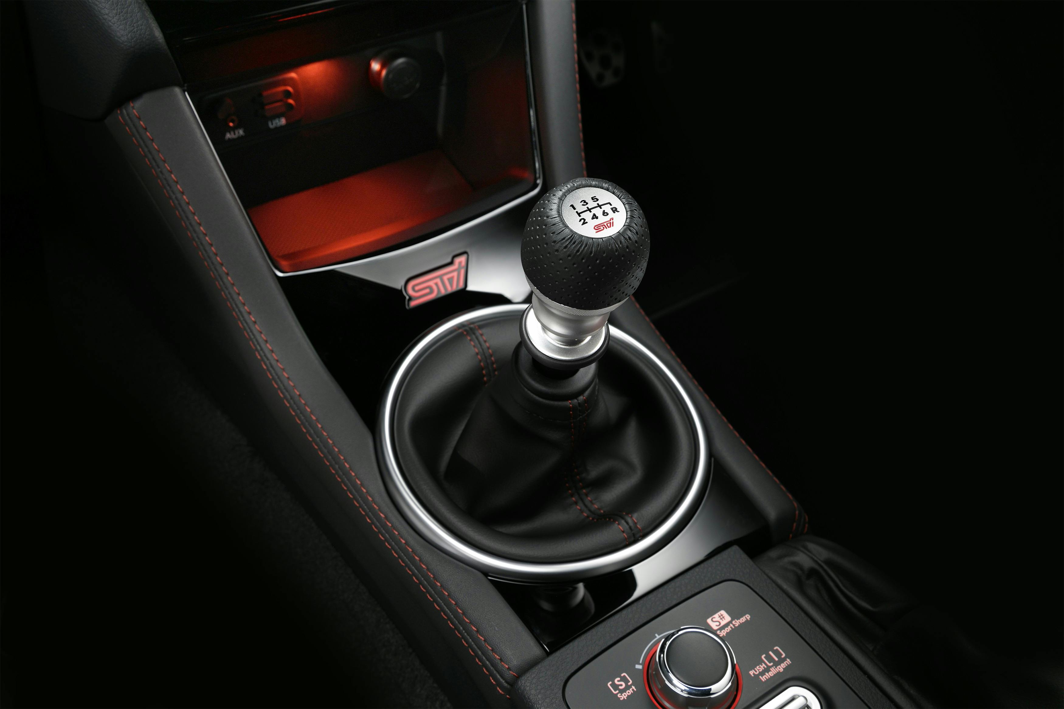Nahaufnahme eines schwarzen Auto-Schaltknüppels mit roter Naht und STI-Logo, umgeben von einer beleuchteten Konsole.