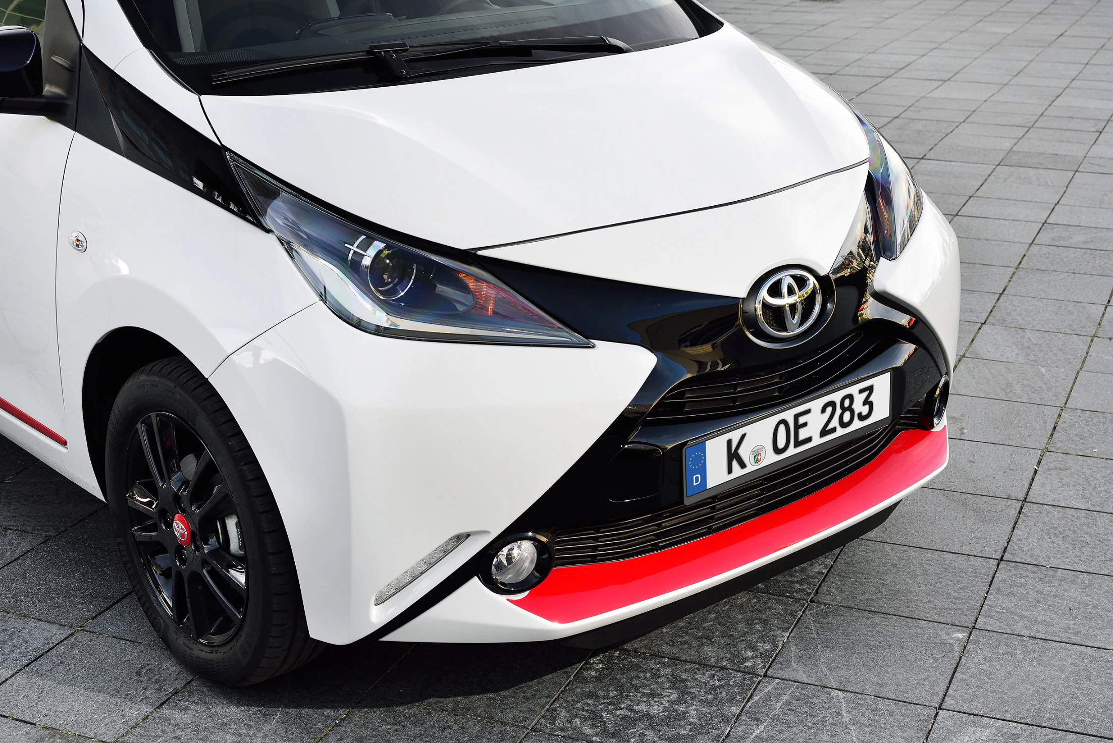 Weißer Toyota Aygo mit schwarzen Akzenten und rotem Streifen an der Stoßstange, fotografiert auf grauem Pflastersteinen.