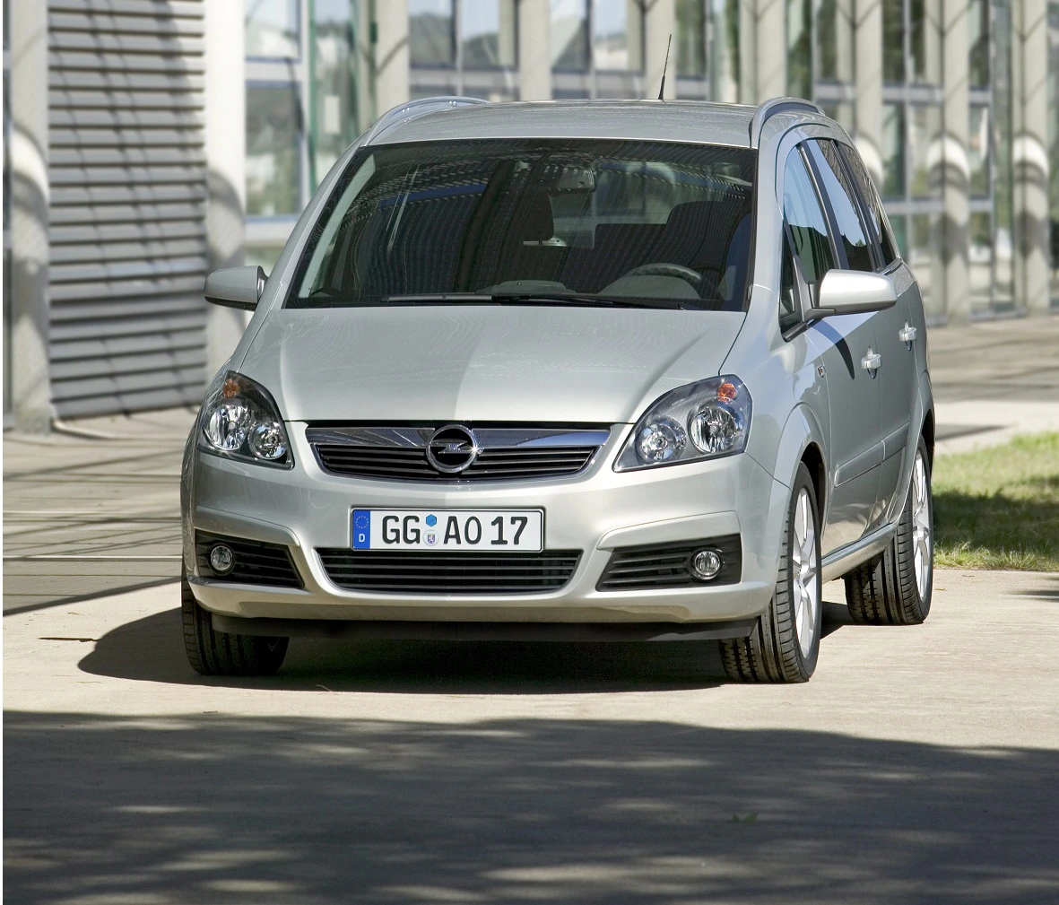 Silberner Opel Zafira steht auf einem Parkplatz vor einem modernen Gebäude. Fahrzeug von vorne betrachtet.