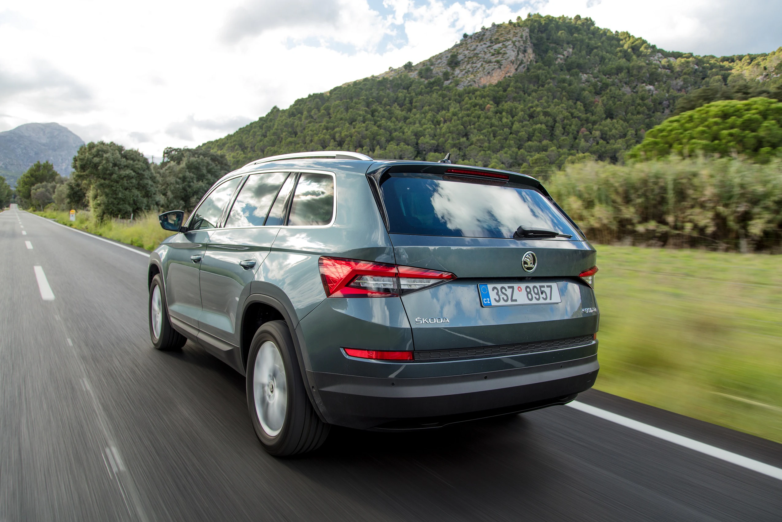 SUV fährt auf einer Landstraße in einer bergigen Landschaft, umgeben von Bäumen und grünem Bewuchs, bei klarem Himmel.