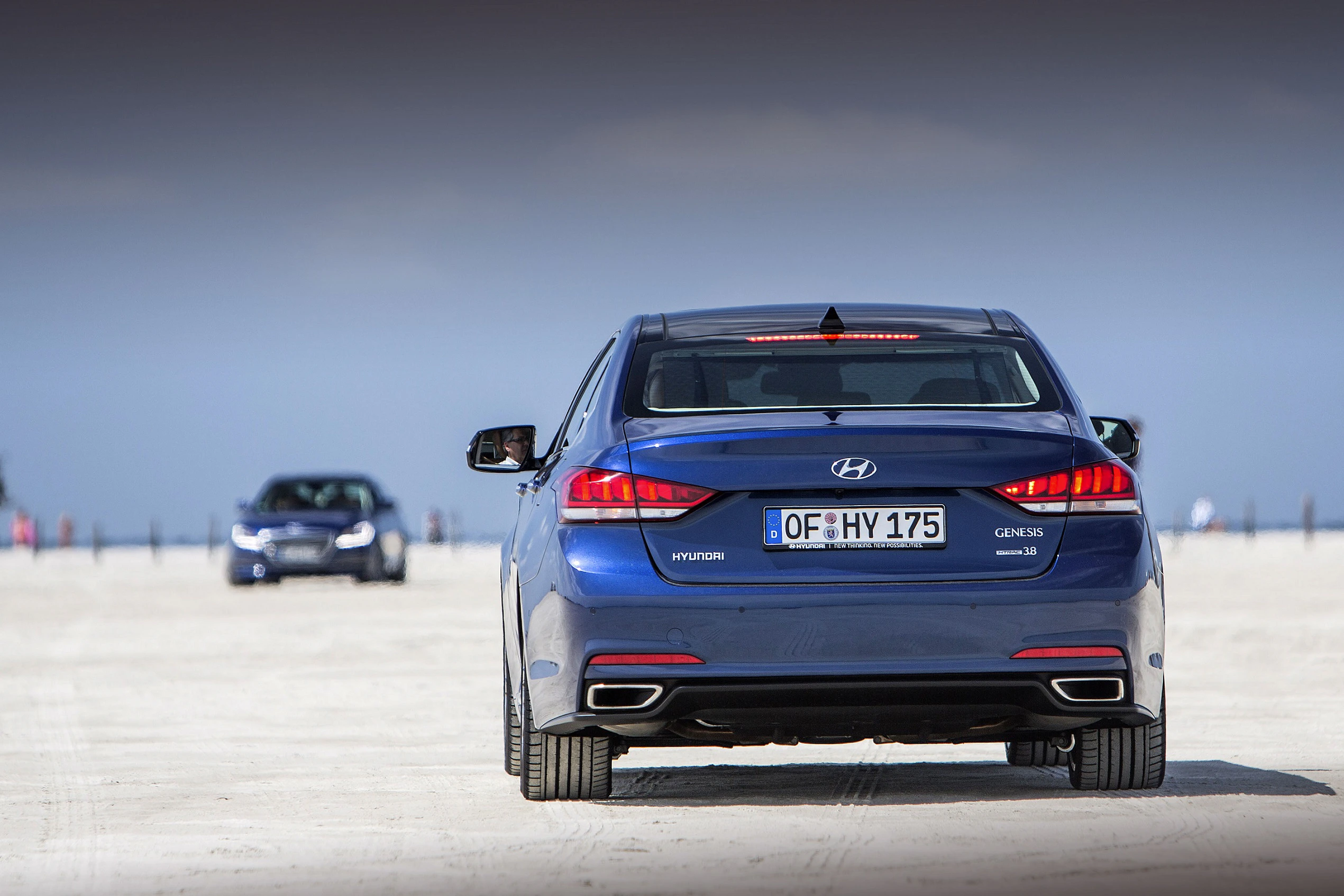 Blaues Hyundai Genesis-Auto fährt auf sandiger Strecke. Im Hintergrund ist ein weiteres Auto sichtbar. Bewölkter Himmel oben.