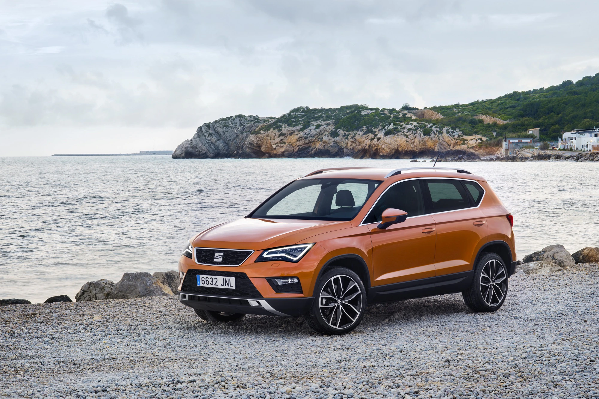 Orangefarbenes SUV parkt auf einem steinigen Küstenabschnitt mit Meer und bewölktem Himmel im Hintergrund. Felsige Landschaft im Hintergrund.