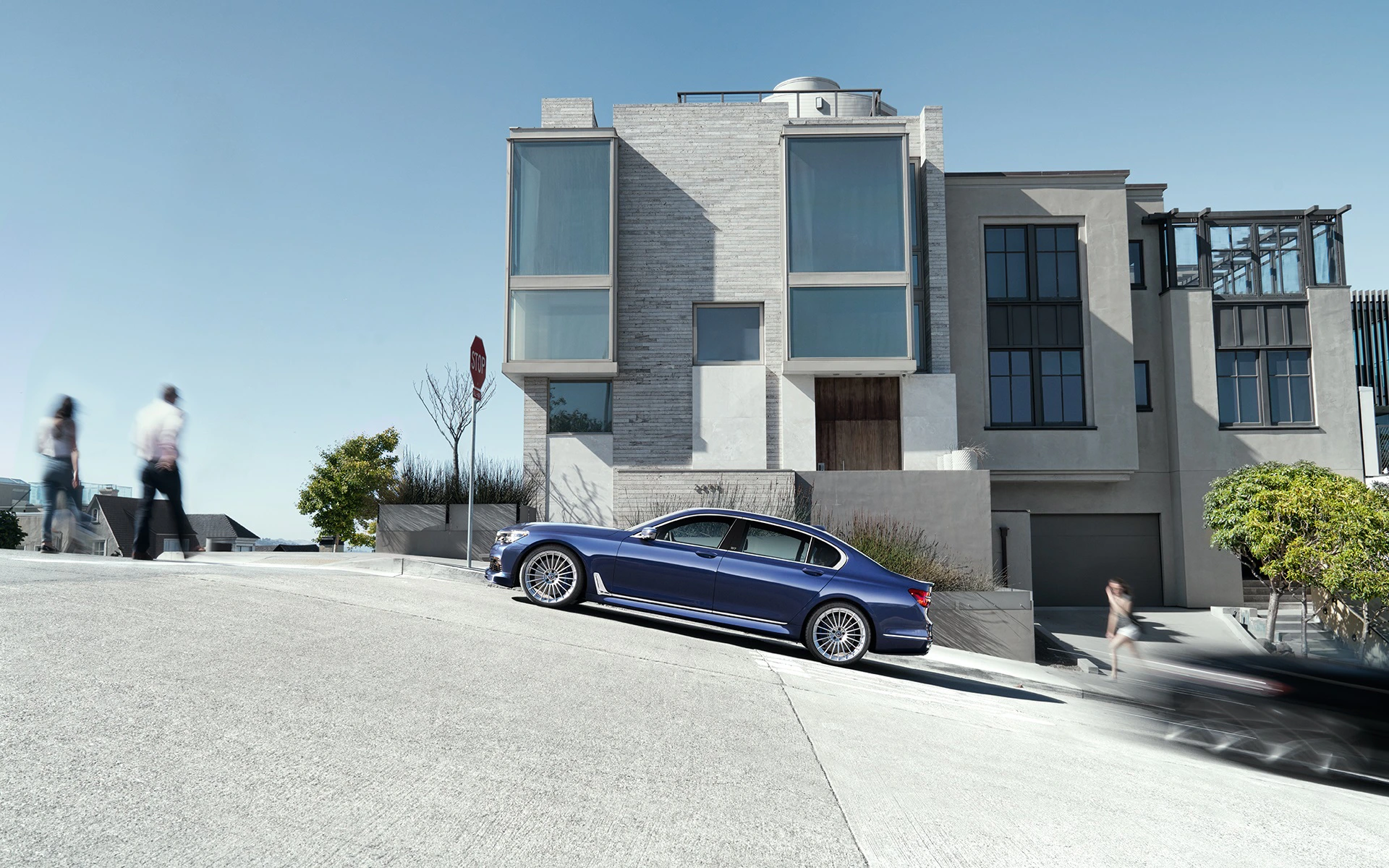 Blaues Auto parkt an steiler Straße vor modernem, mehrstöckigem Haus; Menschen gehen im Hintergrund vorbei.