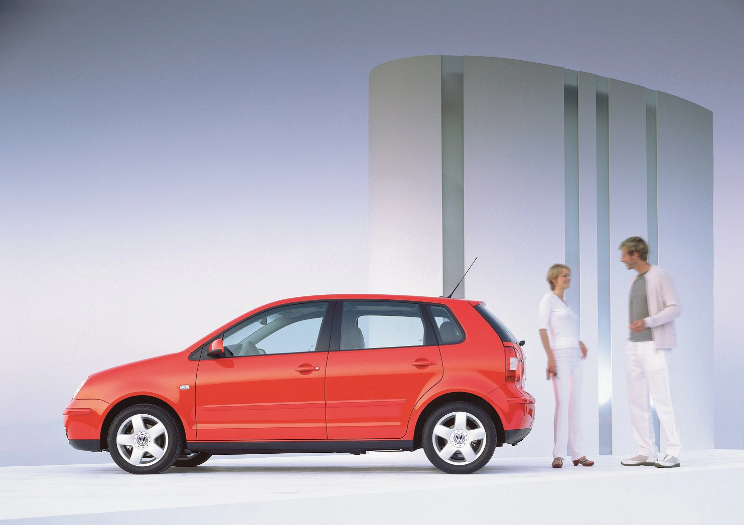 Rotes Auto mit fünf Türen auf weißem Boden vor futuristischer Kulisse; zwei Personen in weißen Outfits stehen rechts und unterhalten sich.