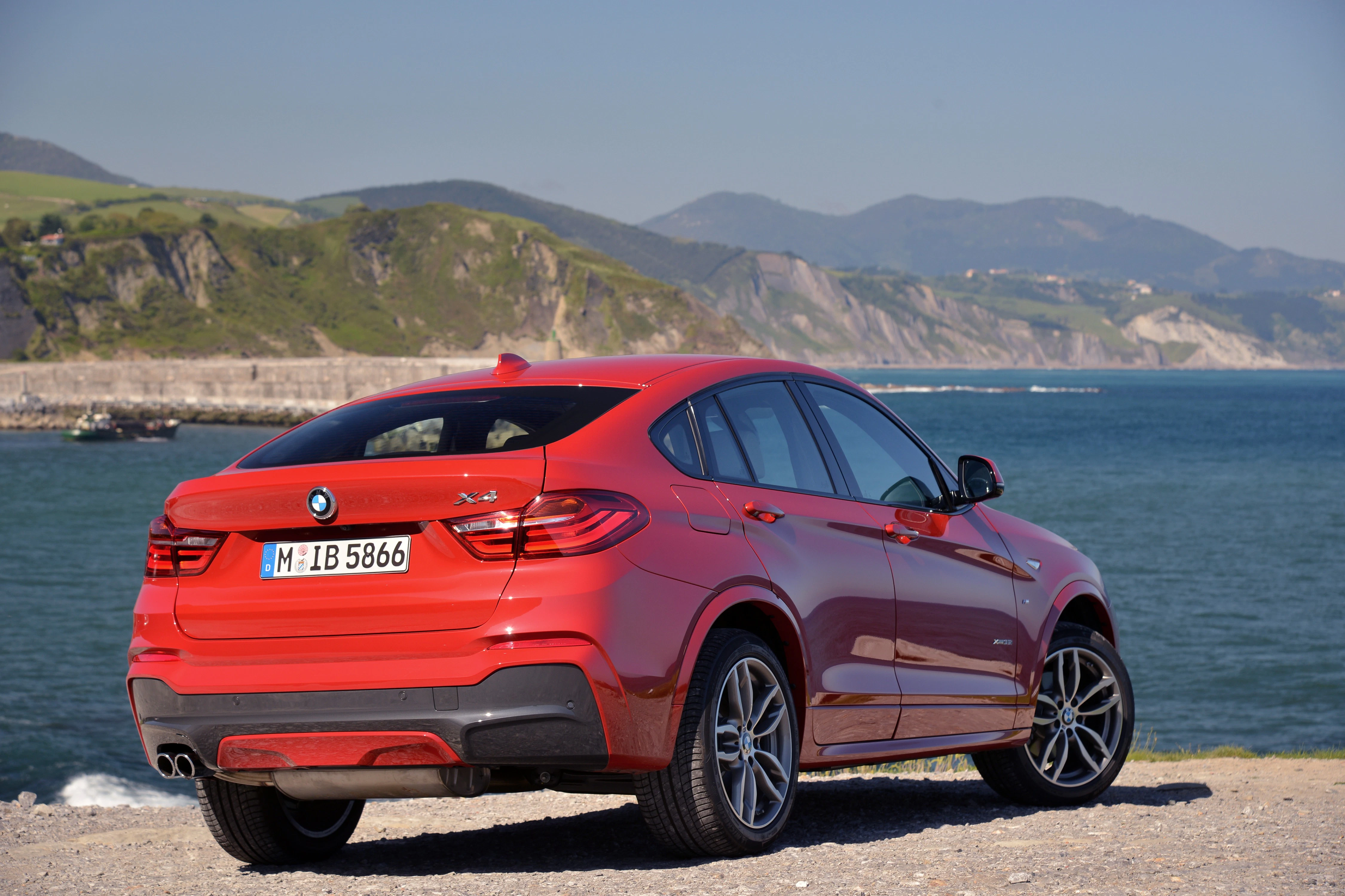 Roter BMW X4 mit Küstenlandschaft im Hintergrund. Auto steht auf einem felsigen Weg, Berge und Meer sind sichtbar unter blauem Himmel.