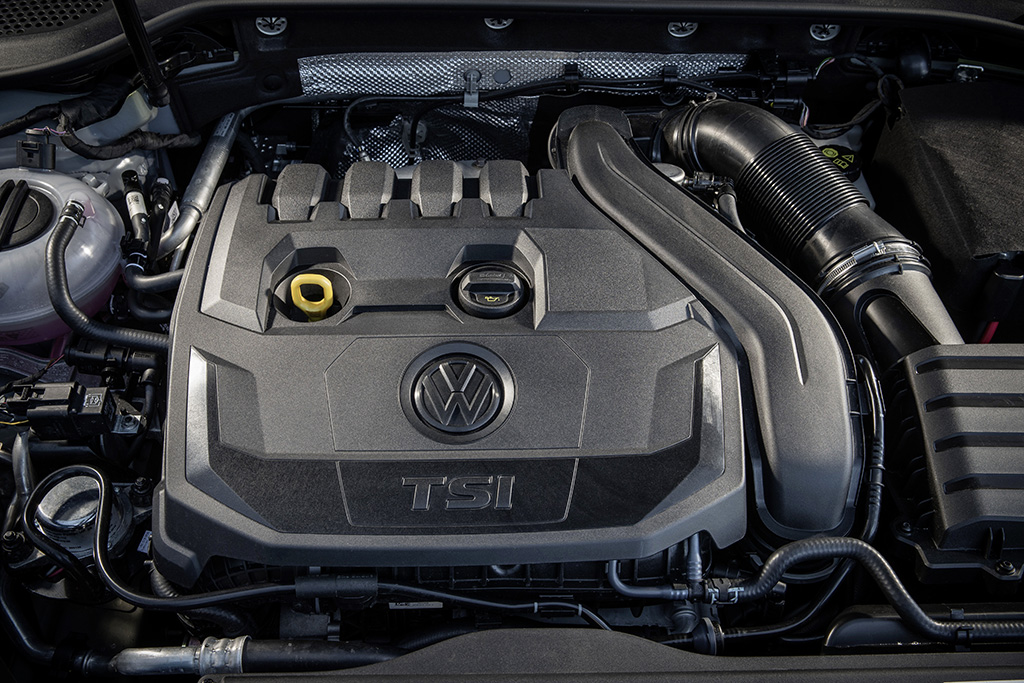 Motorraum mit einem TSI-Motor von Volkswagen, detailreiche Ansicht mit sichtbarem VW-Logo auf der Motorabdeckung, umgeben von Schläuchen und Teilen.
