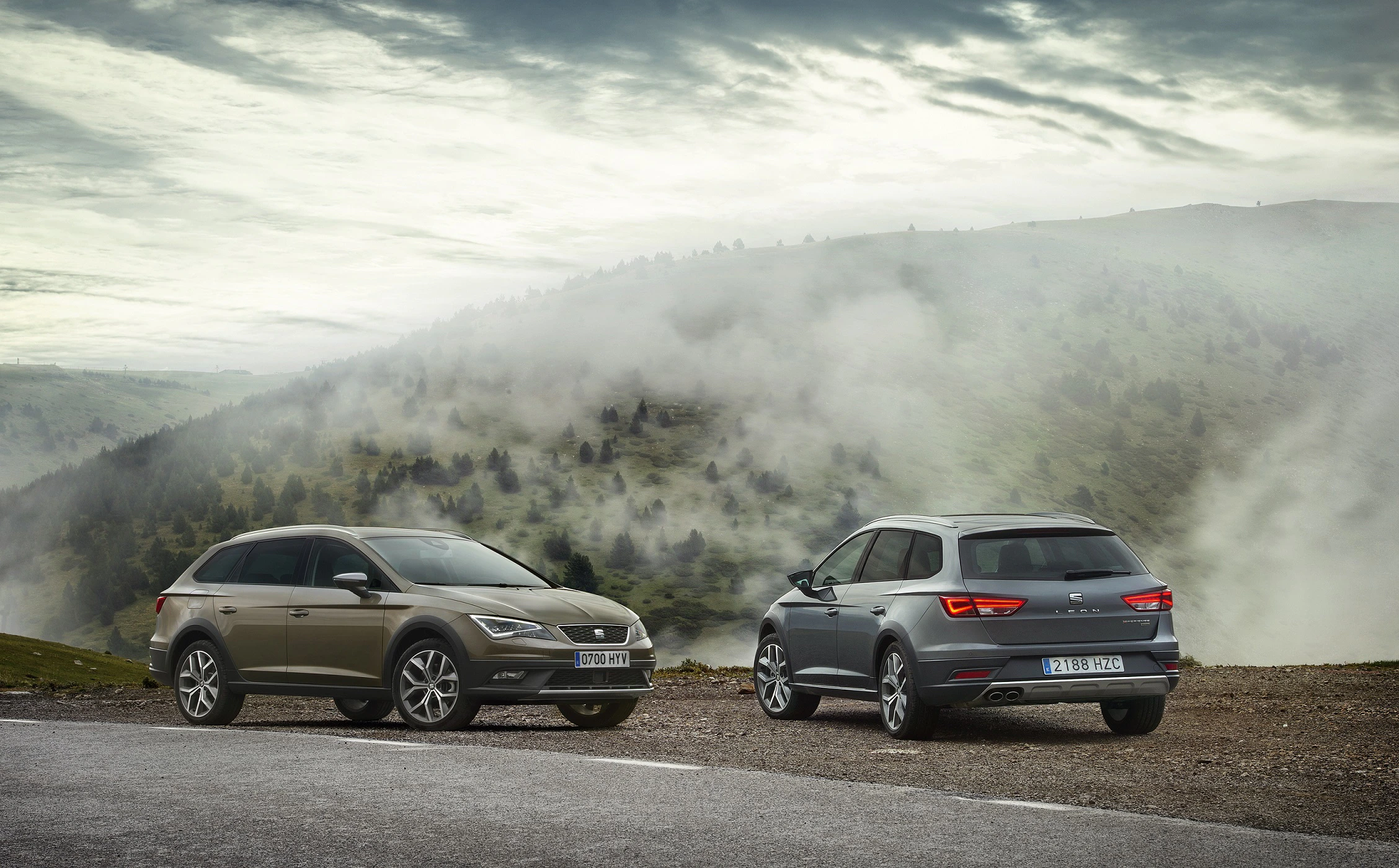 Zwei Autos stehen auf einer Bergstraße mit nebliger Landschaft im Hintergrund.