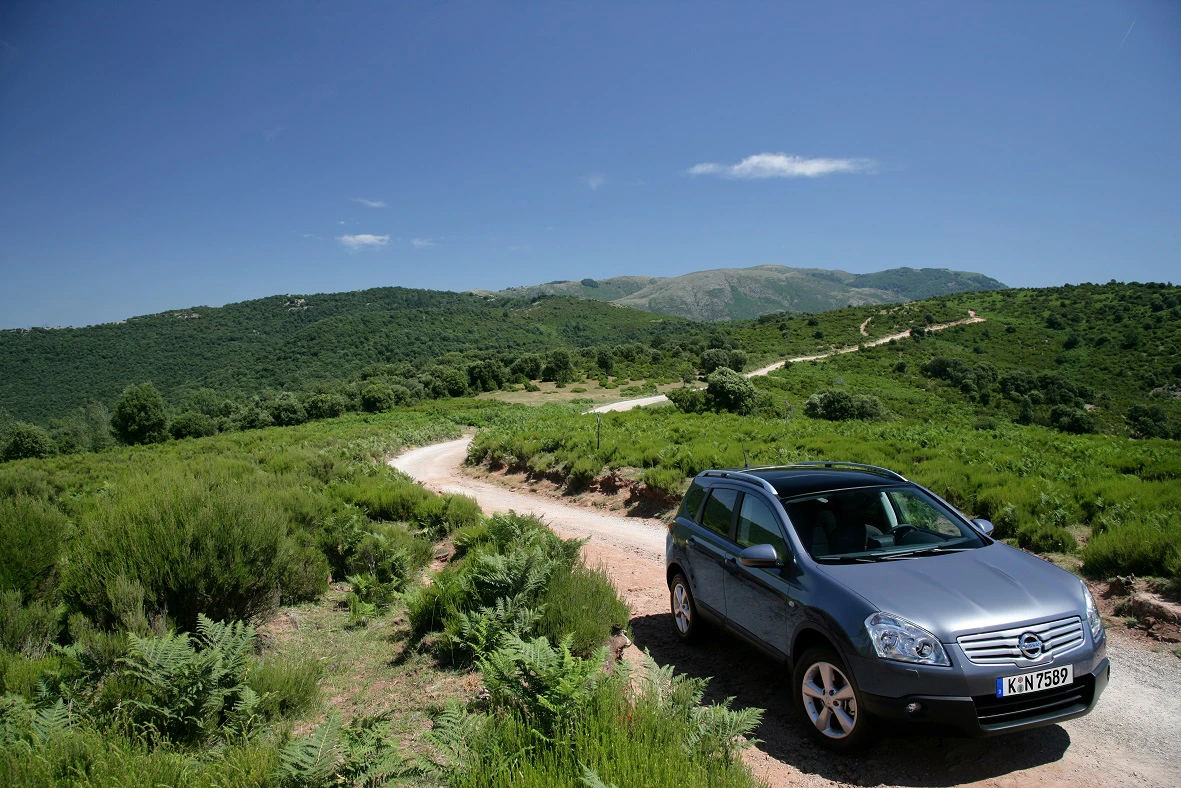 Silberner SUV auf einem kurvigen Landweg in einer grünen, hügeligen Landschaft unter klarem, blauem Himmel.