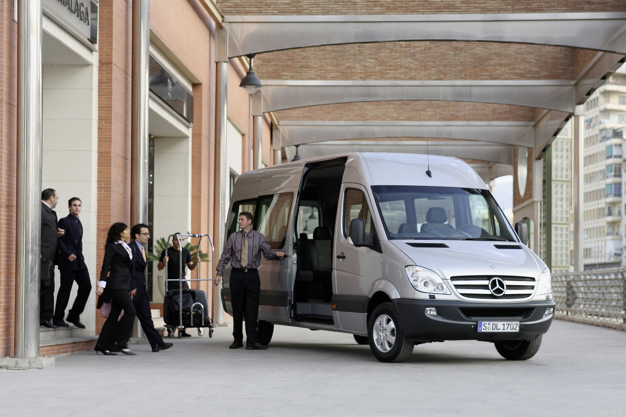 Personengruppe steht neben einem geparkten silbernen Mercedes-Transporter vor einem Gebäude. Einige tragen Uniformen, ein Mann lehnt an der Tür.