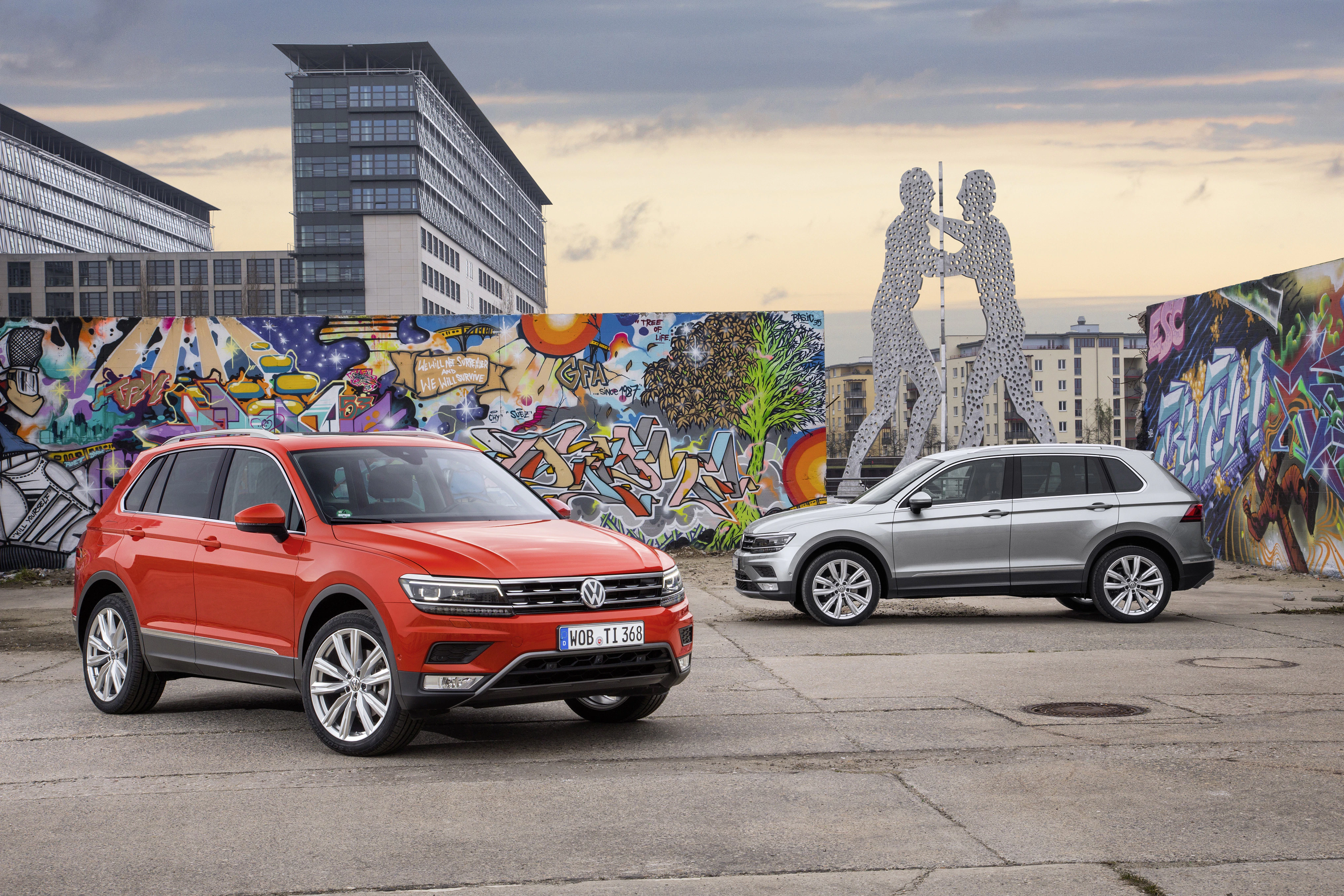 Zwei Volkswagen SUVs stehen auf einem Platz mit Graffiti-Wänden und einer modernen Skulptur im Hintergrund bei bewölktem Himmel.