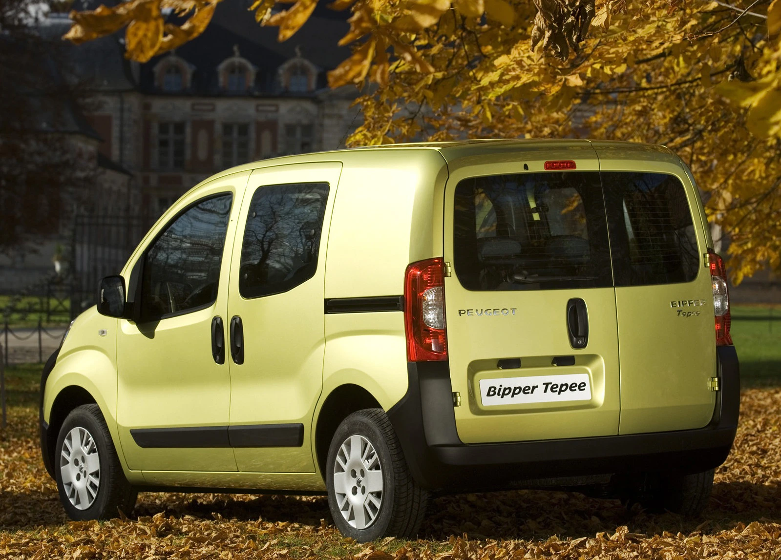Ein gelber Peugeot Bipper Tepee steht auf einem mit Herbstlaub bedeckten Boden, umgeben von Bäumen mit gelbem Laub. Im Hintergrund ist ein Gebäude zu sehen.