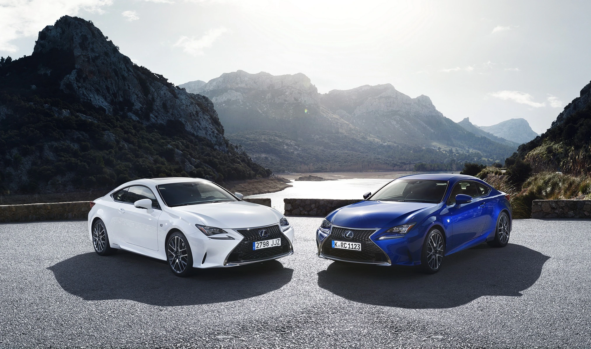 Zwei sportliche Autos, ein weißes und ein blaues, parken auf einer malerischen Straße vor einer beeindruckenden Bergkulisse.