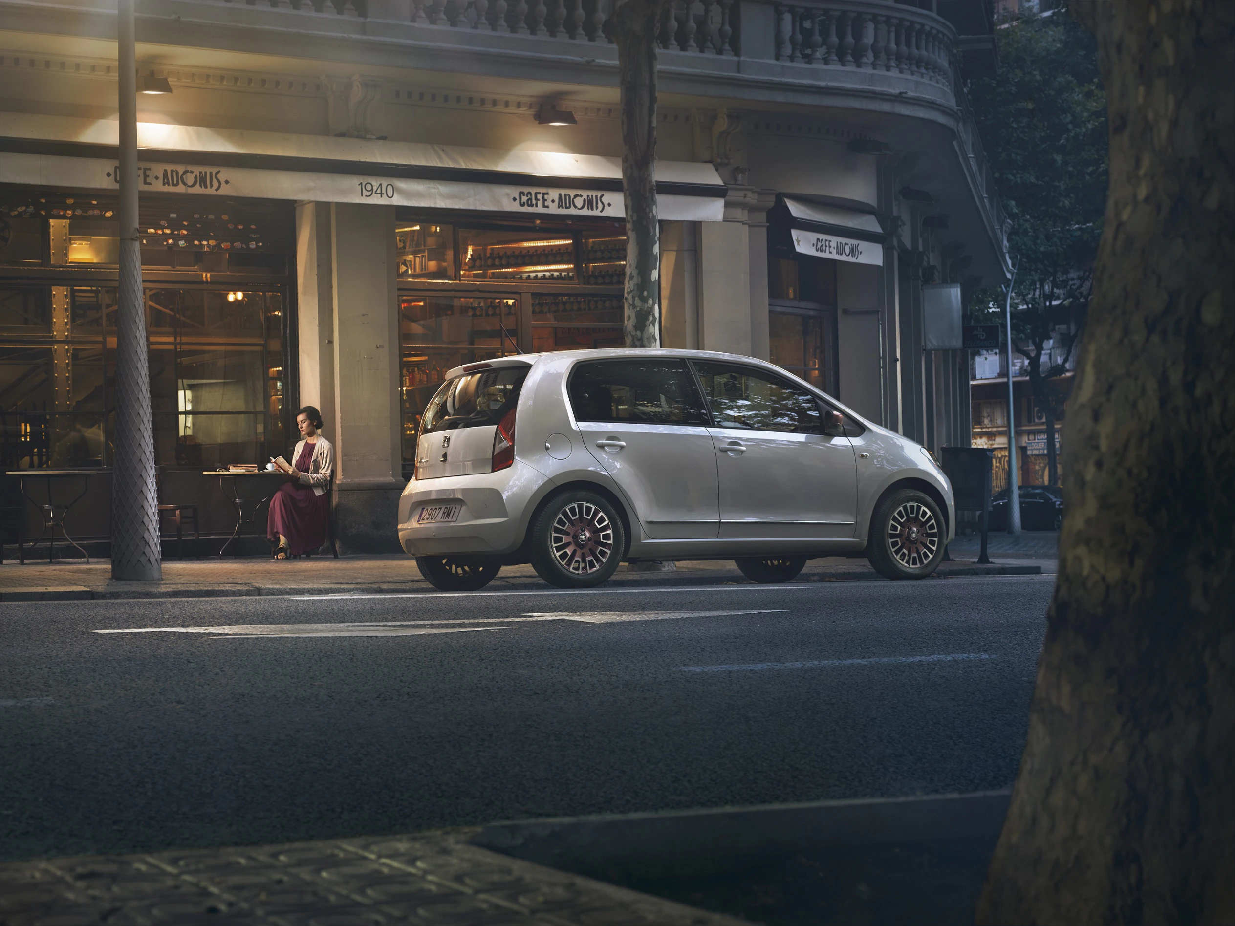 Ein silbernes Auto parkt abends vor einem Café mit beleuchteter Fassade. Zwei Personen sitzen draußen unter einem Baum.