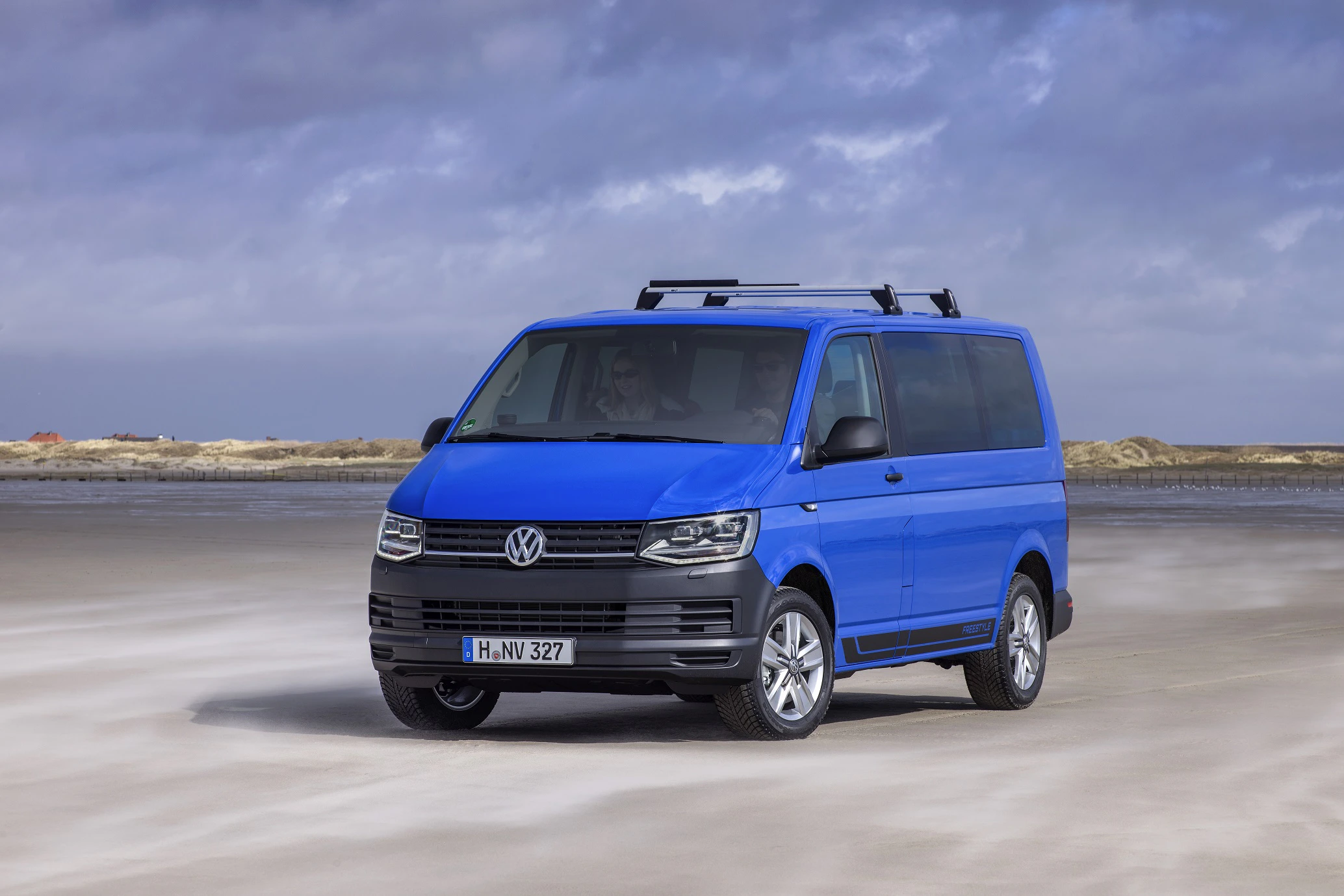 Blauer Volkswagen Transporter fährt an einem sandigen Strand entlang, mit bewölktem Himmel im Hintergrund.