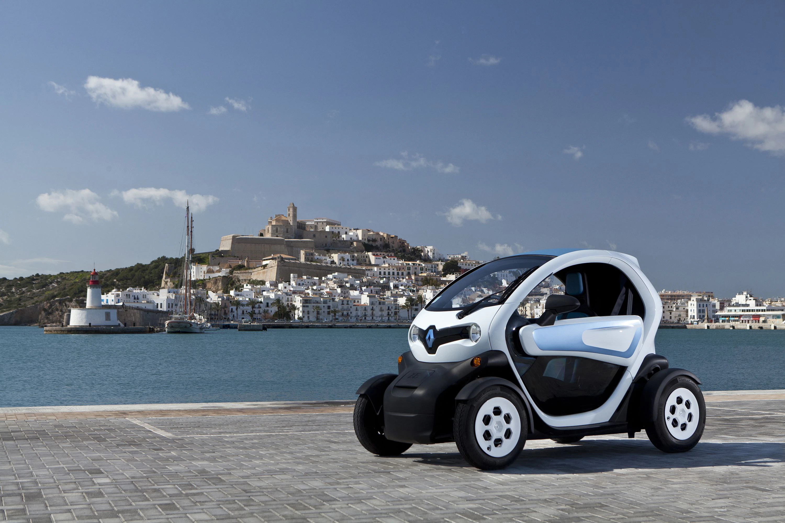 Kompaktes Elektrofahrzeug am Hafen, mit Blick auf eine historische Stadt und das Meer im Hintergrund, unter einem klaren Himmel.