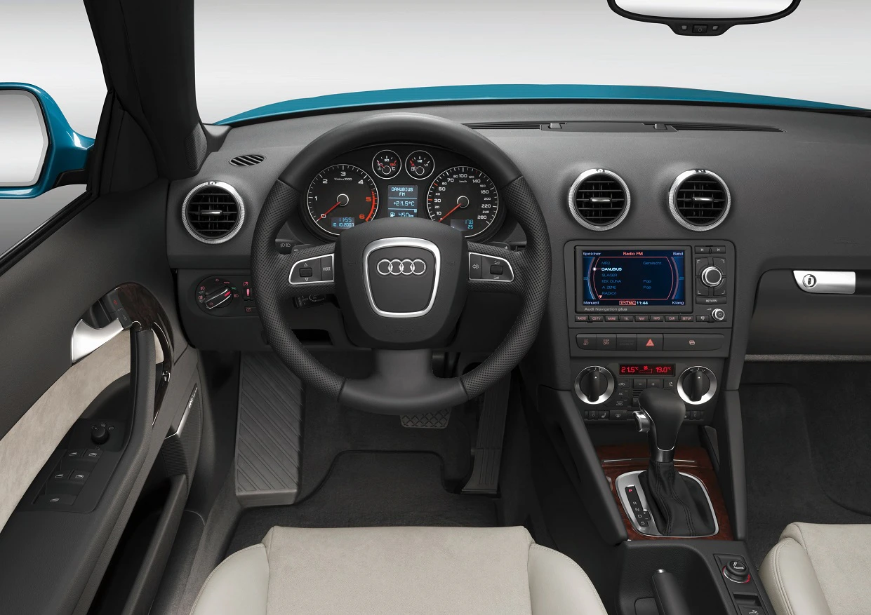 Innenraum eines Audi-Autos mit Lenkrad, Tachometer, Mittelkonsole und einem Infotainment-Bildschirm auf einem schwarzen Armaturenbrett.