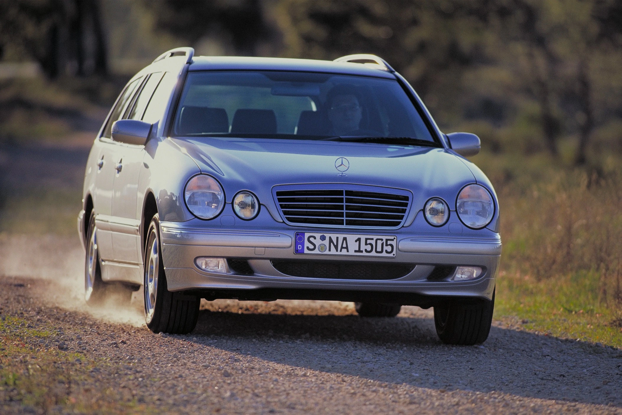 Silberner Kombi Mercedes-Benz E-Klasse fährt auf einem Schotterweg in einer ländlichen Umgebung, Bäume im Hintergrund.