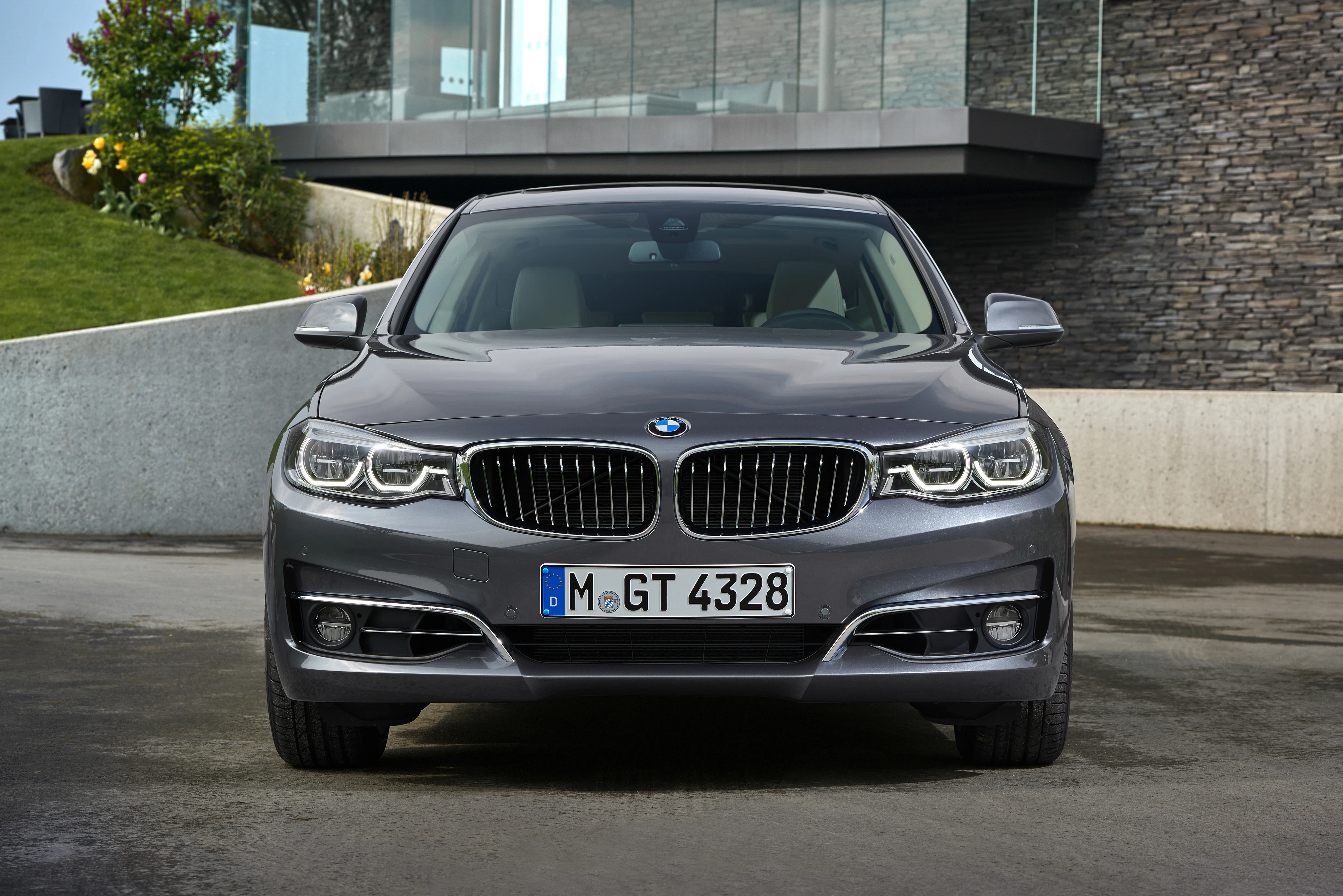 Grauer BMW auf nasser Straße vor modernem Gebäude, Ansicht von vorne. Pflanzen und Steinwand sichtbar. Nummernschild: M-GT 4328.