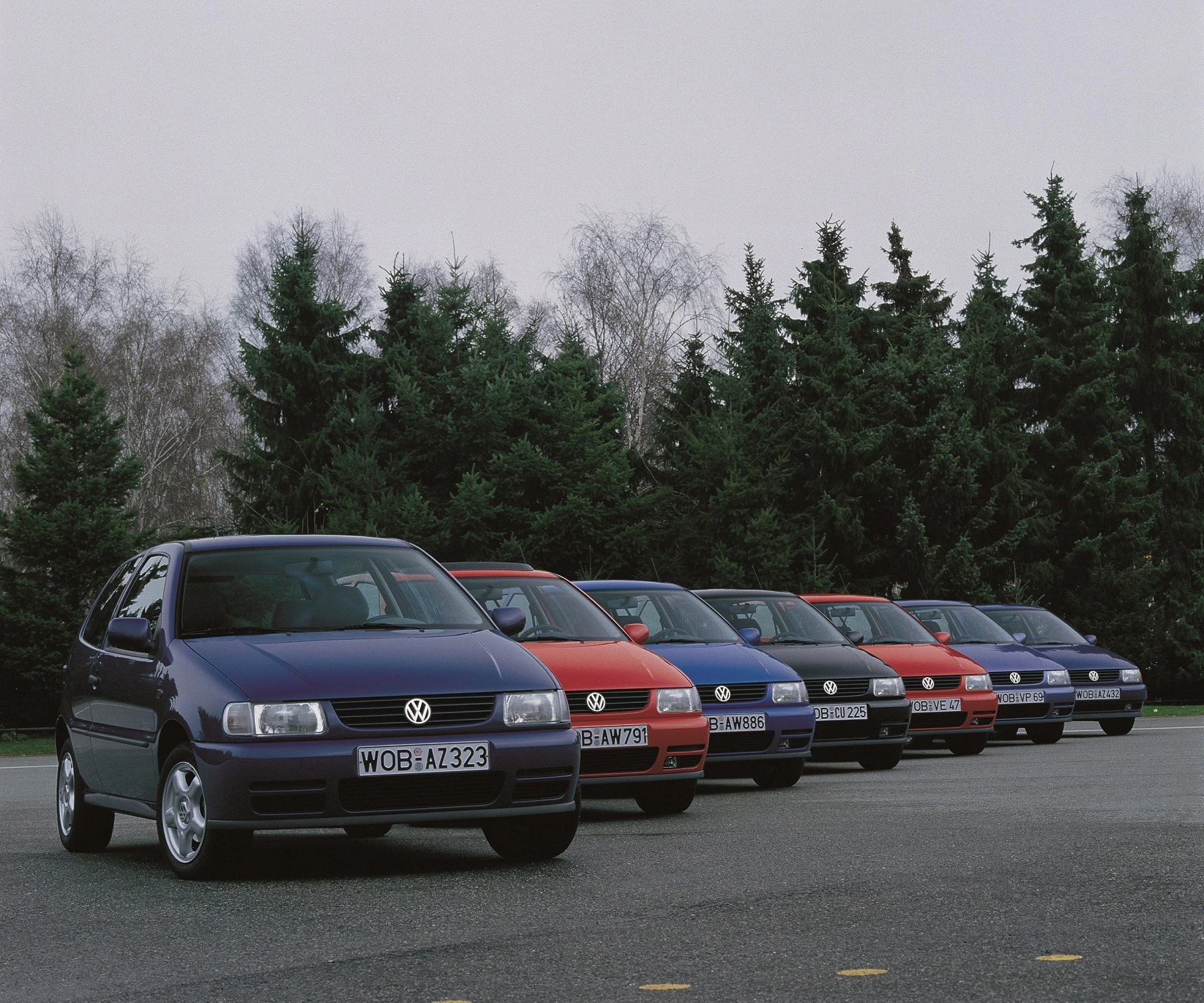 Sechs verschiedenfarbige Volkswagen Golf-Pkws stehen nebeneinander auf einer Straße, umgeben von Bäumen im Hintergrund.