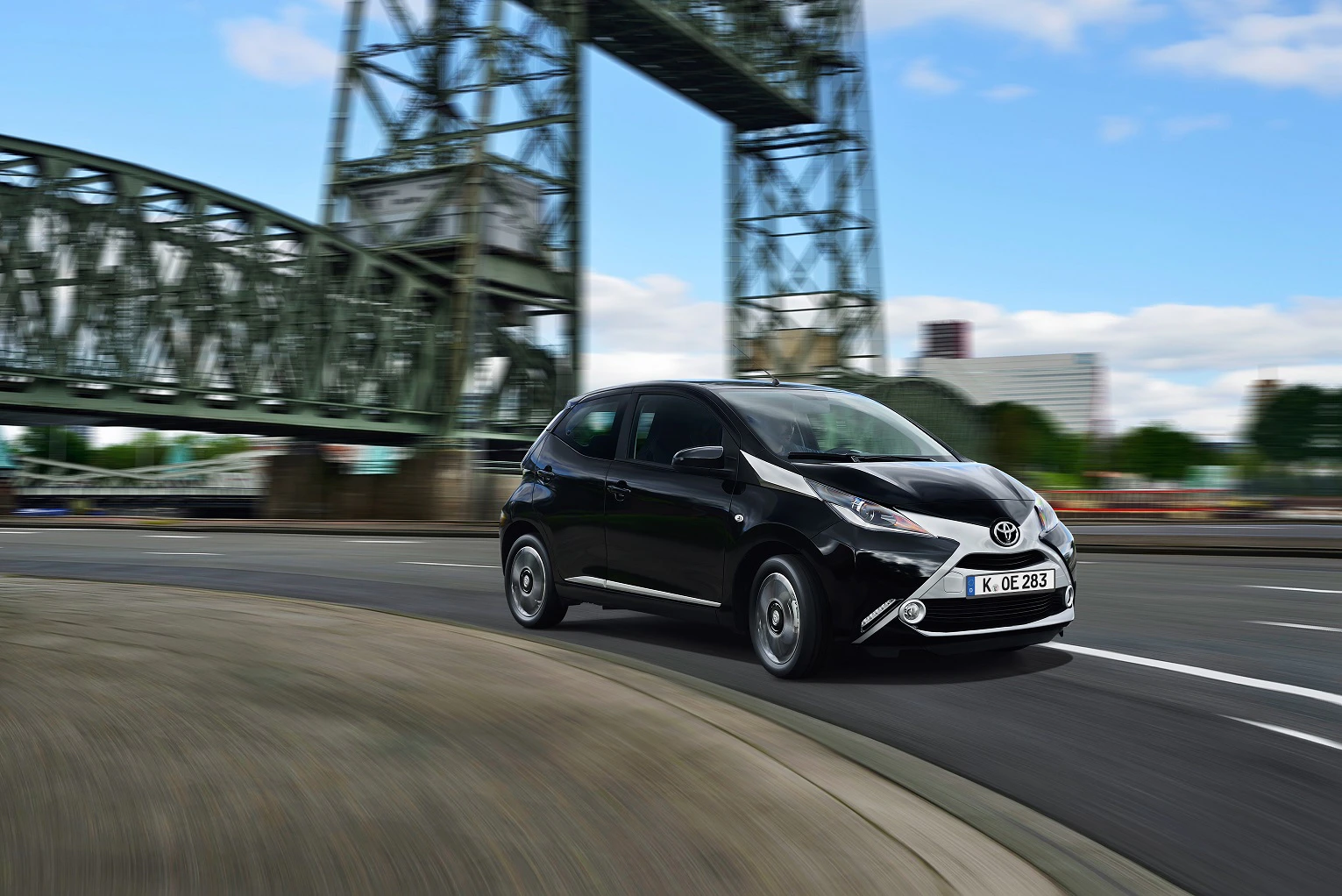 Schwarzer Toyota Aygo fährt auf Stadtstraße unter Brücke, bewölkter Himmel im Hintergrund, leichter Bewegungseffekt sichtbar.