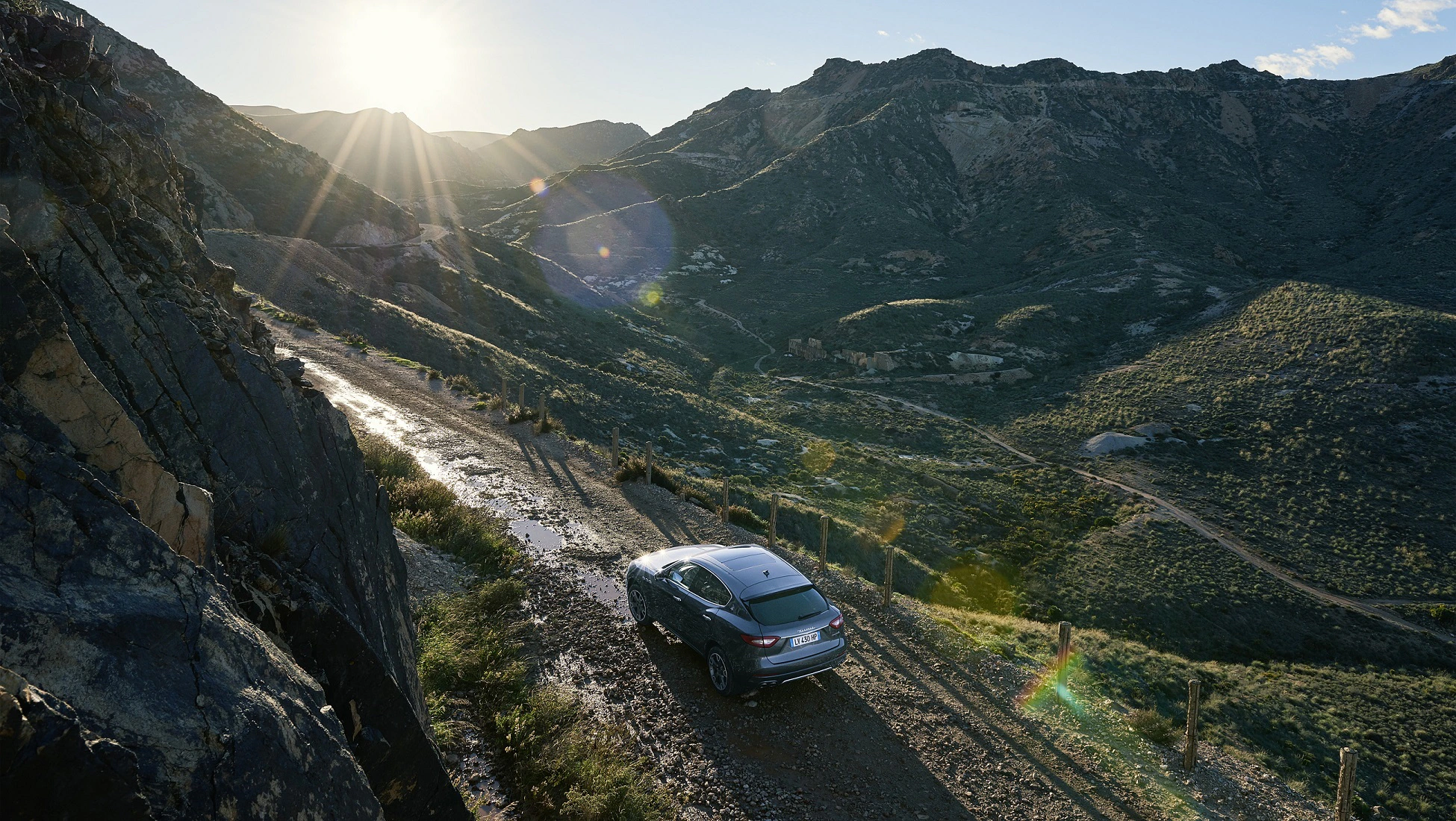 Ein Auto fährt auf einem steinigen Bergweg, umgeben von grünen Hügeln und Felsen, bei tief stehender Sonne am klaren Himmel.
