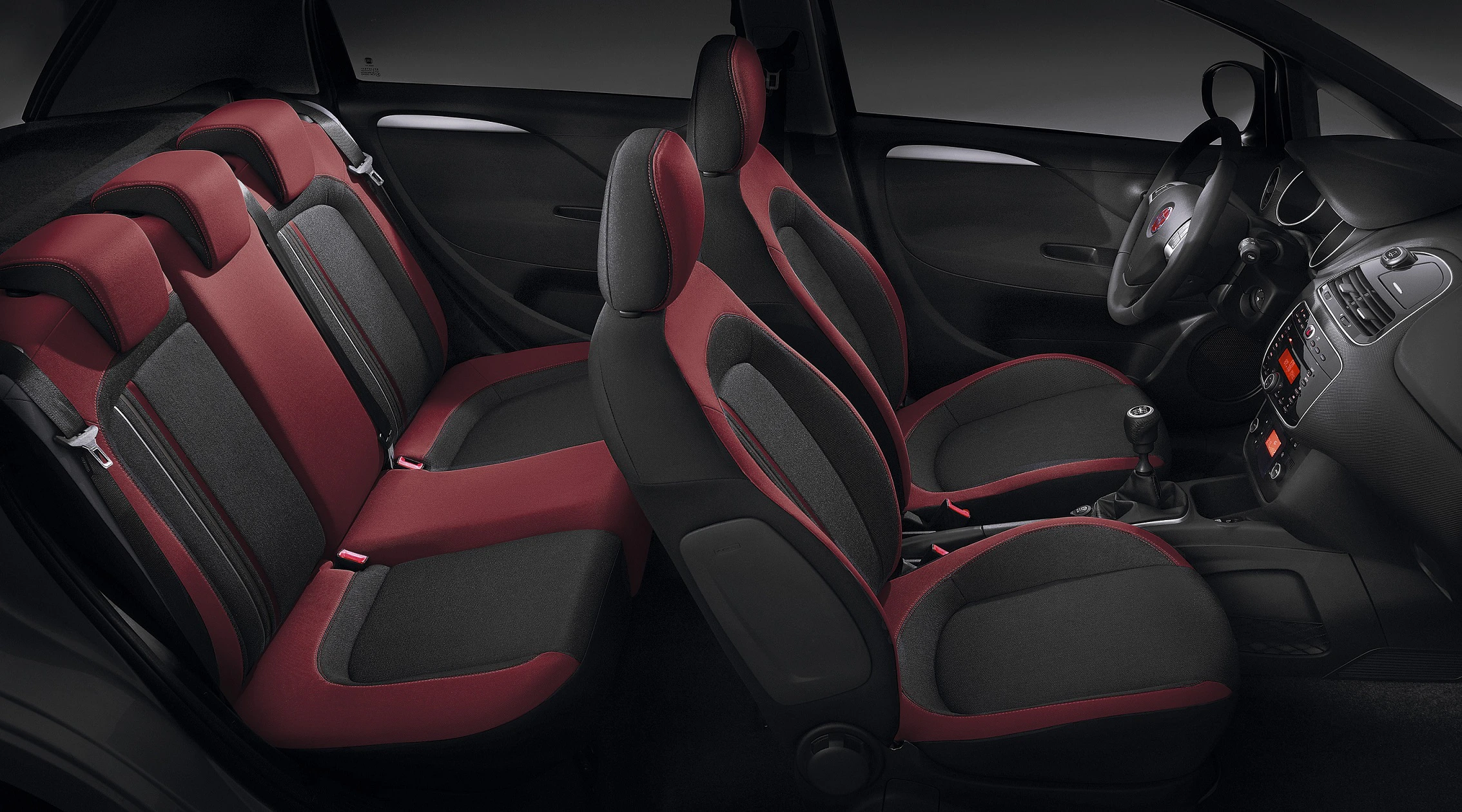 Innenraum eines Autos mit roten und grauen Stoffbezügen, fünf Sitze, moderne Armaturen und manuelles Getriebe.