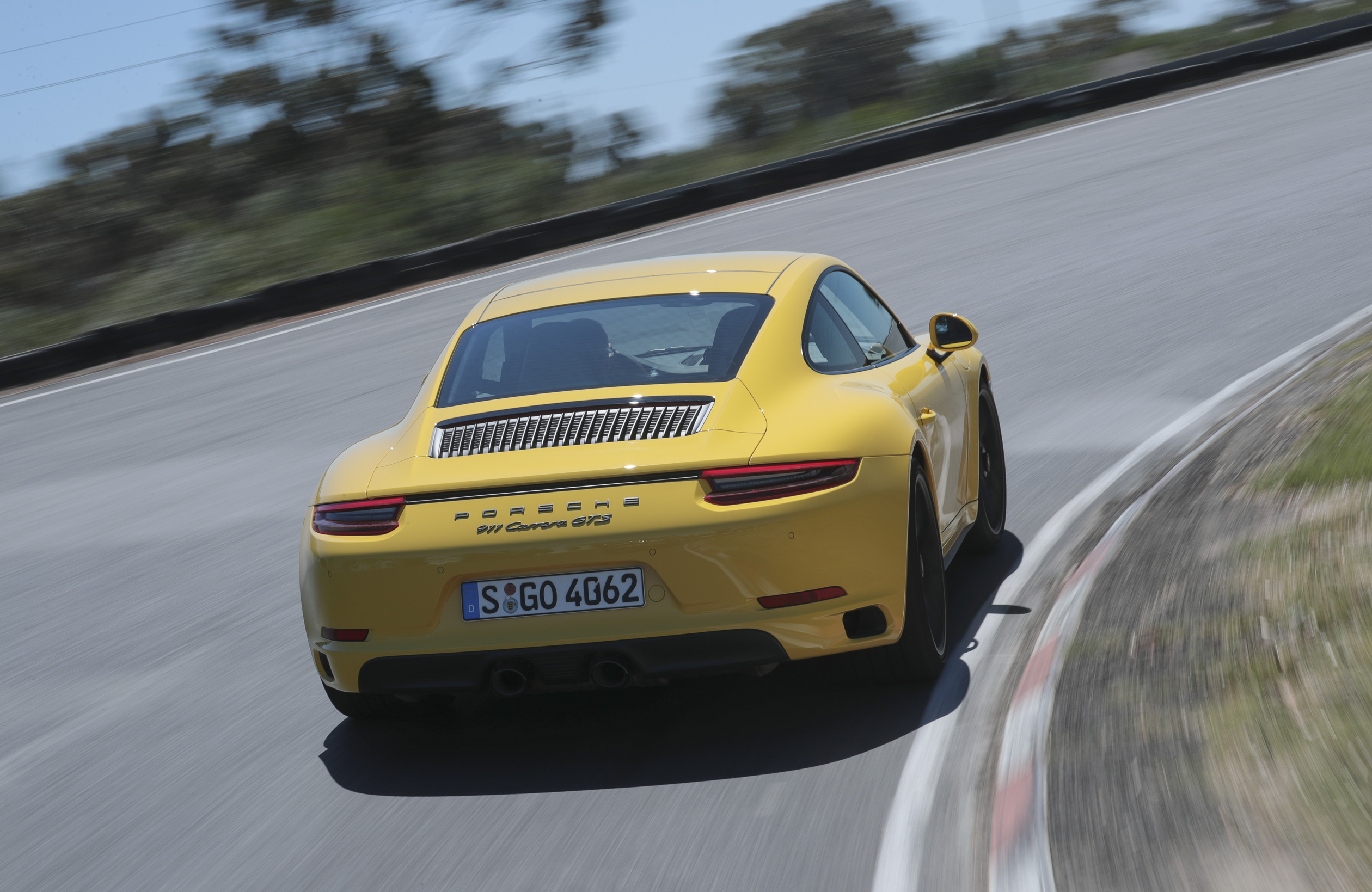 Gelber Sportwagen fährt eine gebogene Rennstrecke entlang. Bäume im Hintergrund, klare Straße, sonniges Wetter.