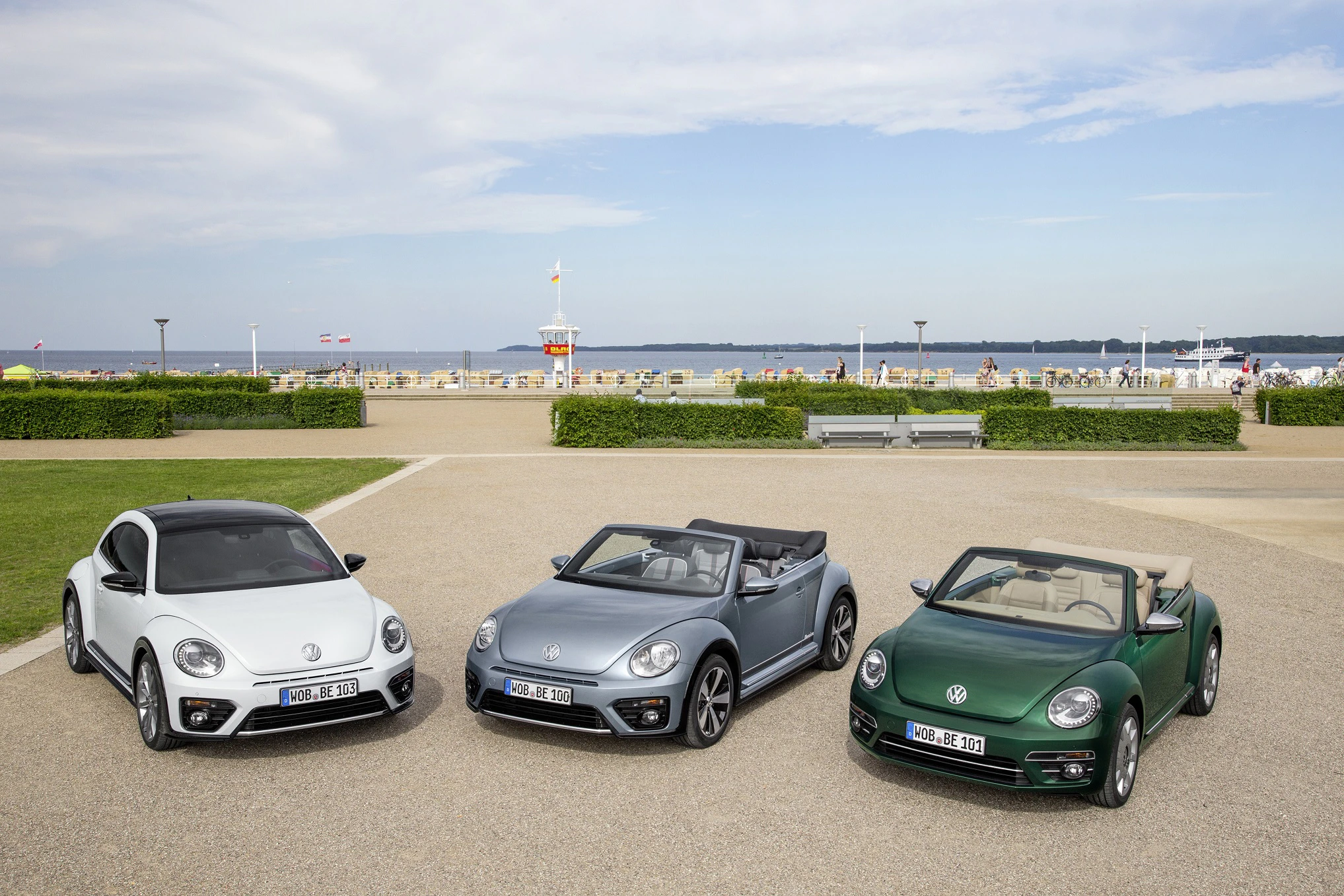 Drei VW Beetle Cabriolets in Weiß, Grau und Grün stehen auf einem Platz vor einer Strandpromenade mit blauen Himmel und Meer im Hintergrund.