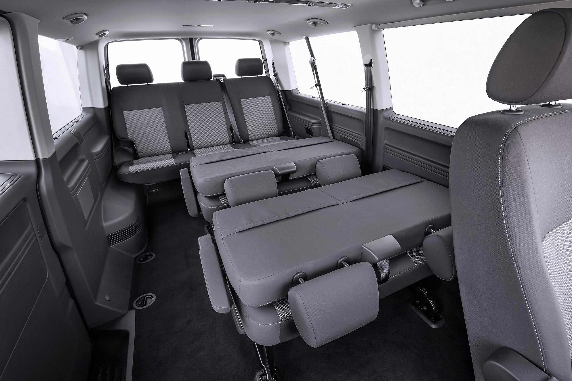 Innenansicht eines geräumigen Transporters mit umgeklappten, grauen Sitzen, die eine Liegefläche bilden. Minimalistisches, modernes Design.