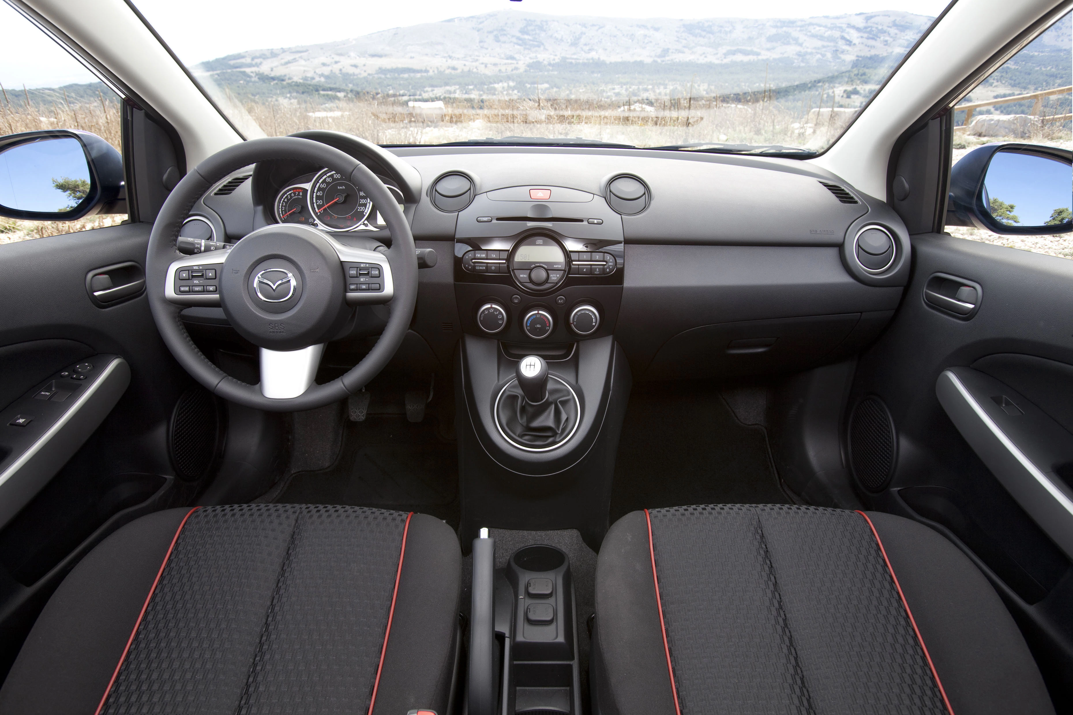 Mazda-Innenraum mit Lenkrad, Armaturenbrett und Schalthebel, schwarzes Design mit roten Nähten, Blick durch die Windschutzscheibe auf Landschaft.
