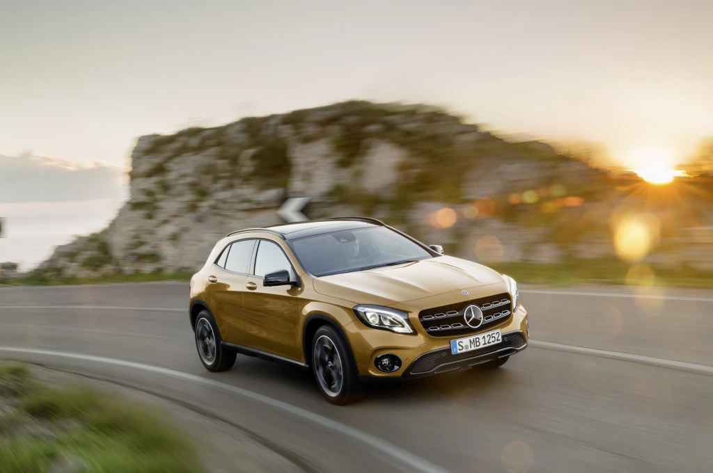 Goldener SUV Mercedes auf kurviger Straße bei Sonnenuntergang, Felsen und Natur im Hintergrund, dynamische Fahrbewegung.