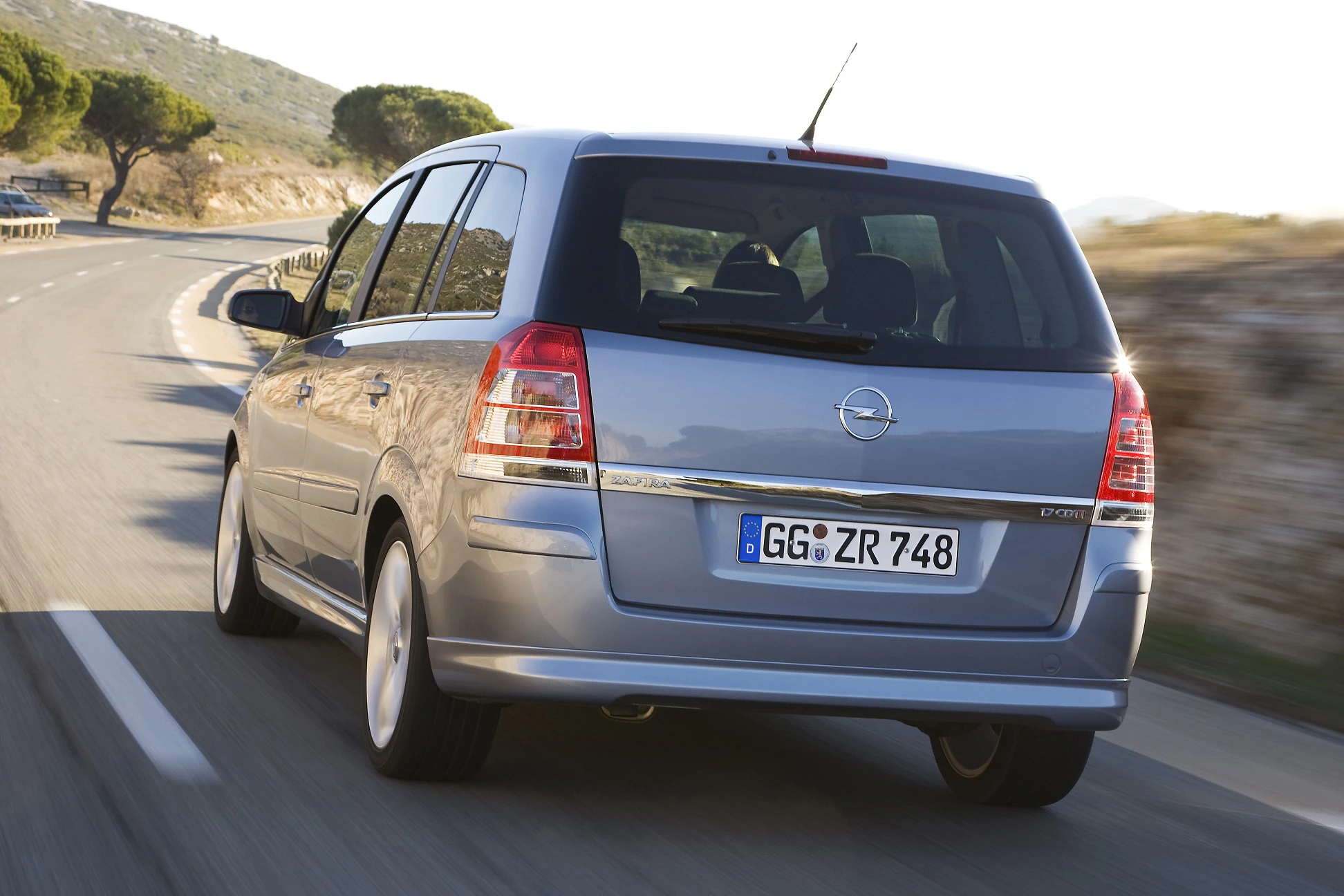 Silberner Opel Zafira fährt auf einer kurvigen Landstraße bei sonnigem Wetter, umgeben von Bäumen und Hügellandschaft.