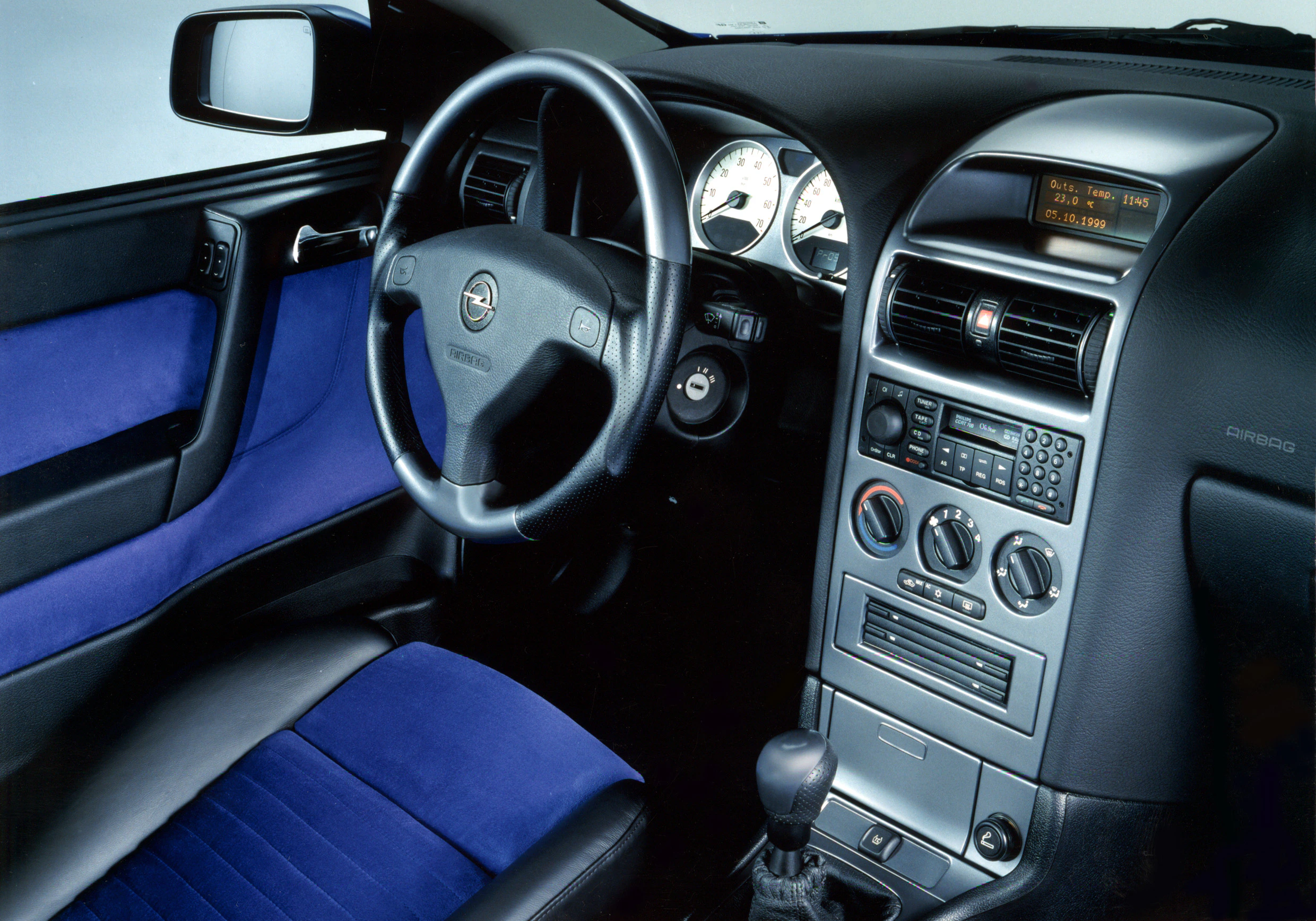 Innenraum eines Autos mit blauer und schwarzer Polsterung, Lenkrad links, Armaturenbrett mit Anzeigen und Bedienelementen.
