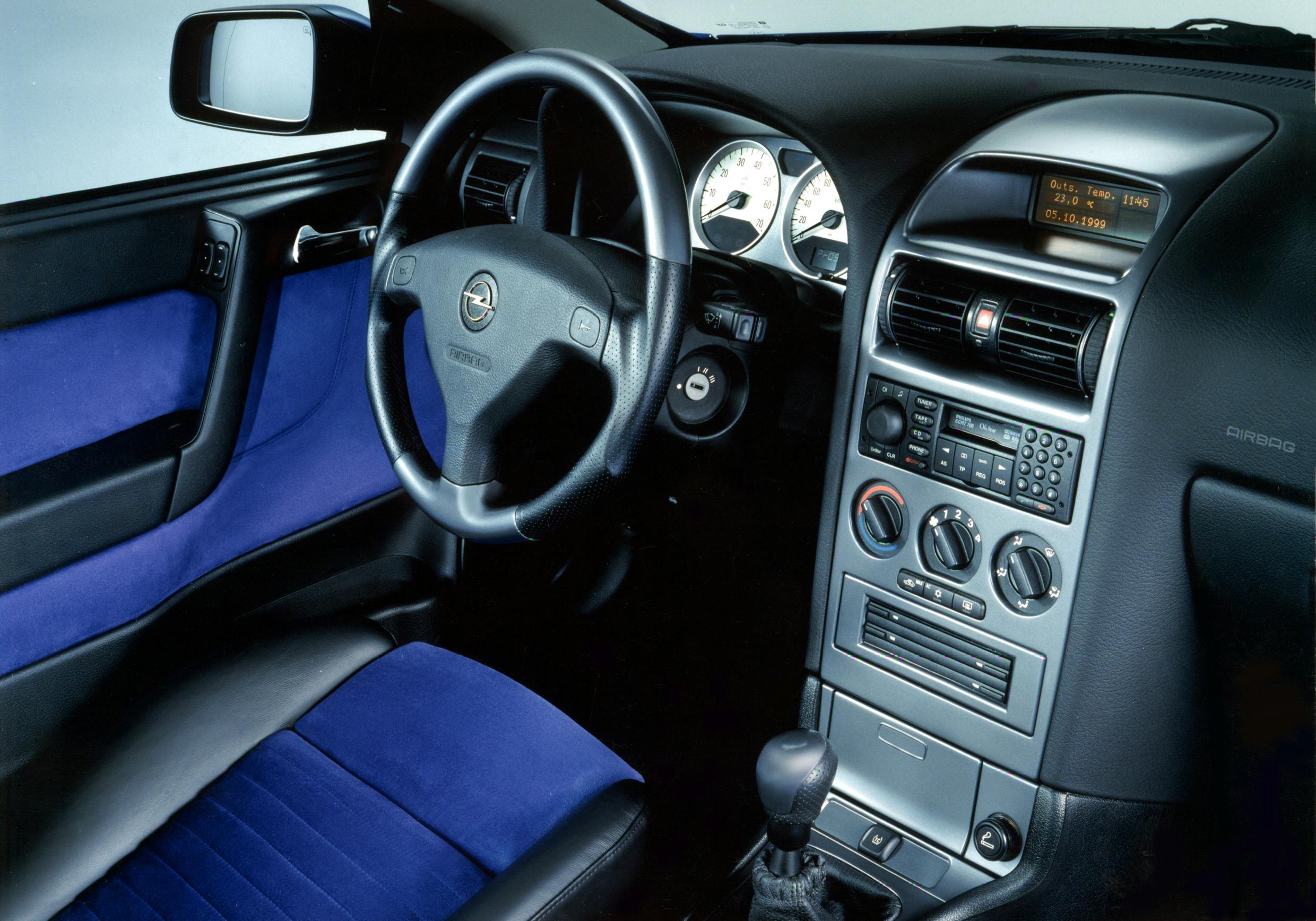 Innenraum eines Autos mit blauer und schwarzer Polsterung, Lenkrad links, Armaturenbrett mit Anzeigen und Bedienelementen.