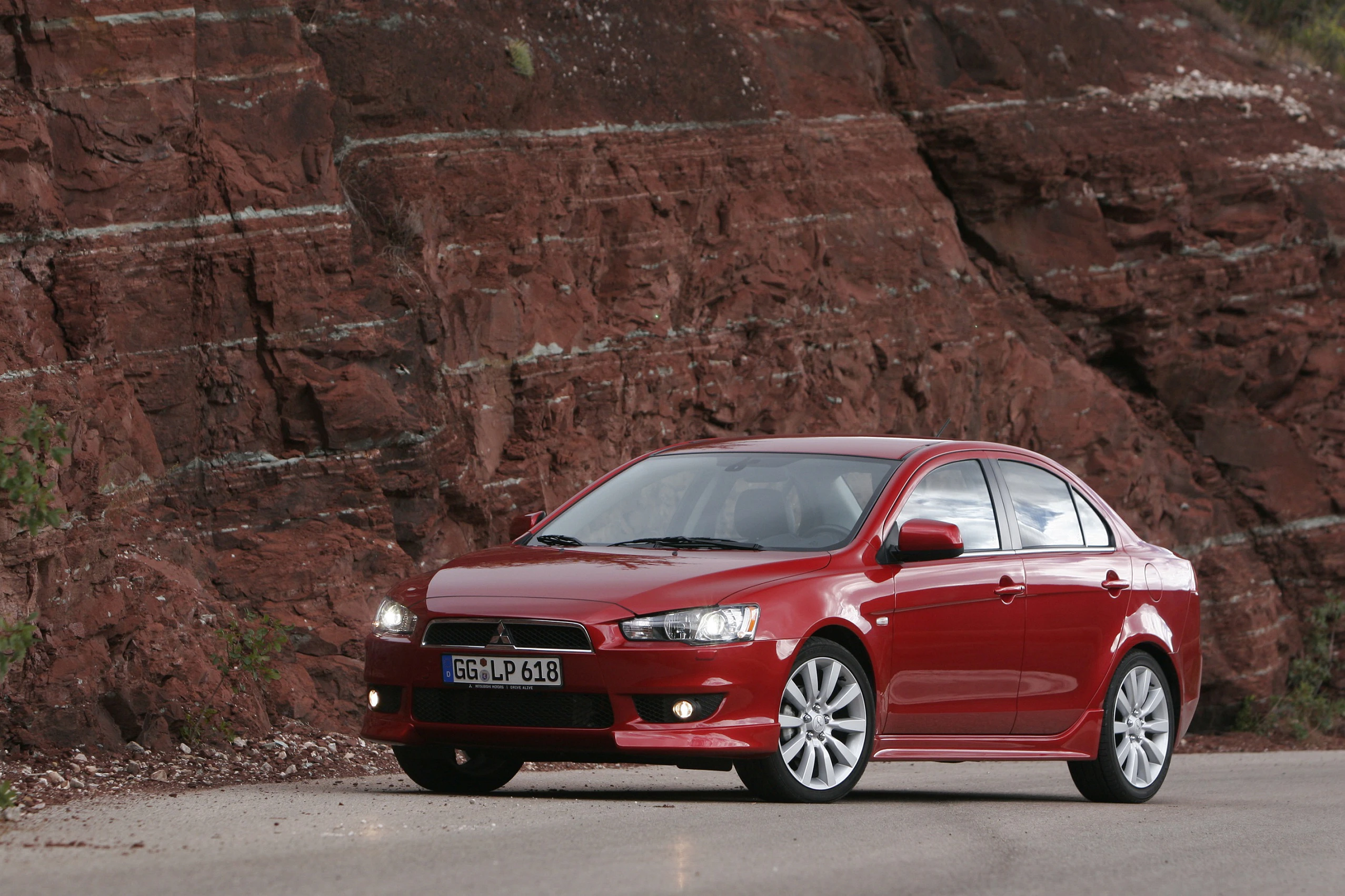 Roter Sportwagen parkt auf einer Straße neben einer steilen, roten Felswand. Das Auto hat silberne Felgen und sportliches Design.