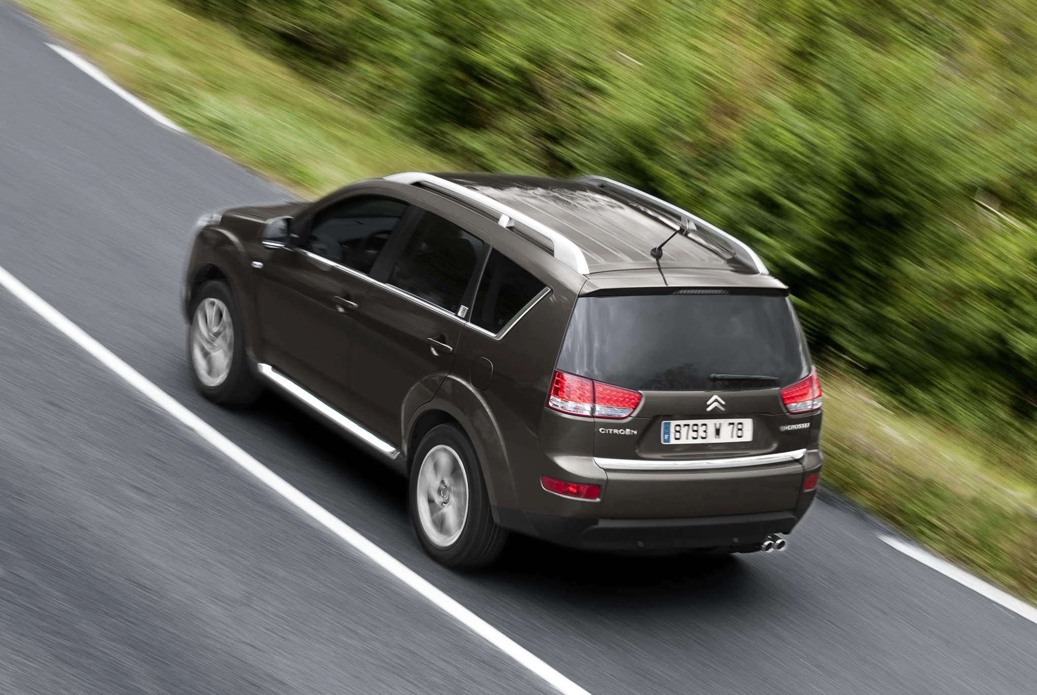 Dunkelgrauer SUV fährt auf einer Landstraße, von oben fotografiert, mit grüner Vegetation am Straßenrand.