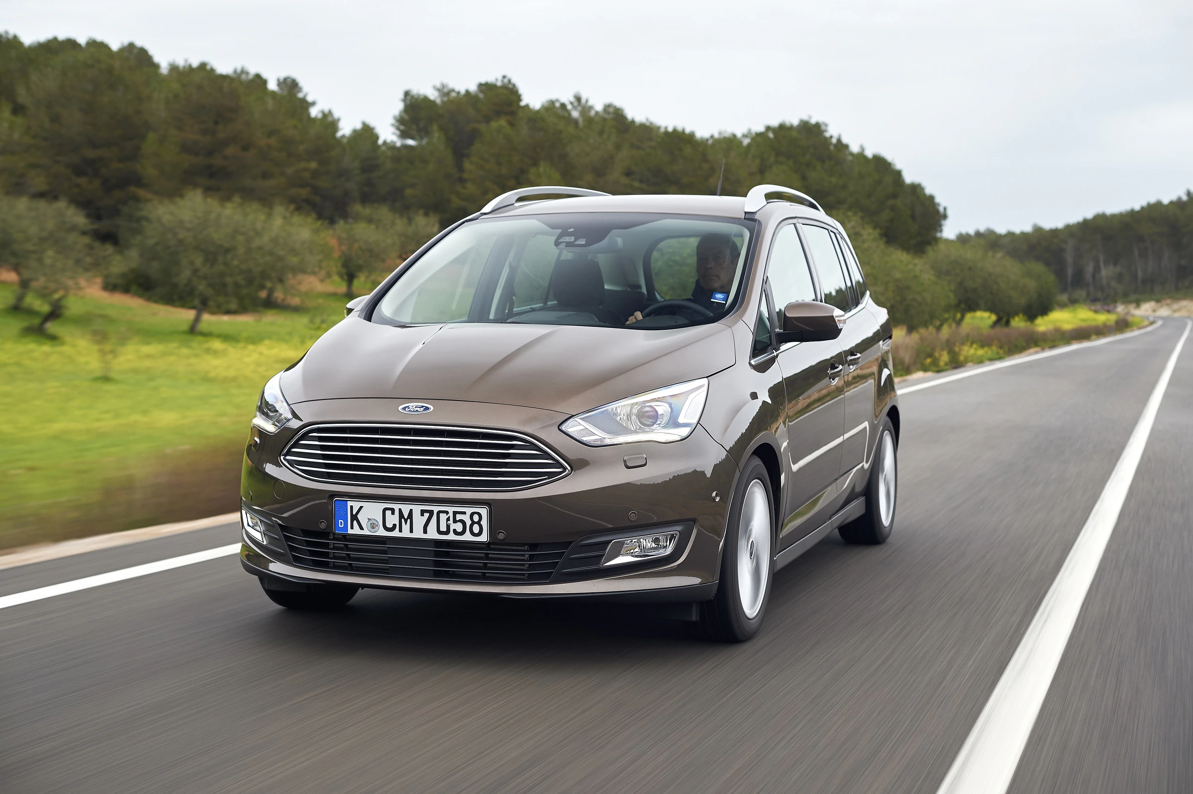 Brauner Ford C-Max fährt auf einer Landstraße entlang grüner Felder und Bäume unter bewölktem Himmel.